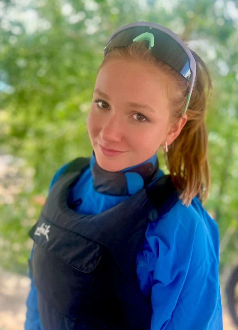 A young woman with light skin, reddish-brown hair tied back, wearing sunglasses on her head, a blue sports jacket, and a black life vest, standing outdoors with blurred green foliage in the background.