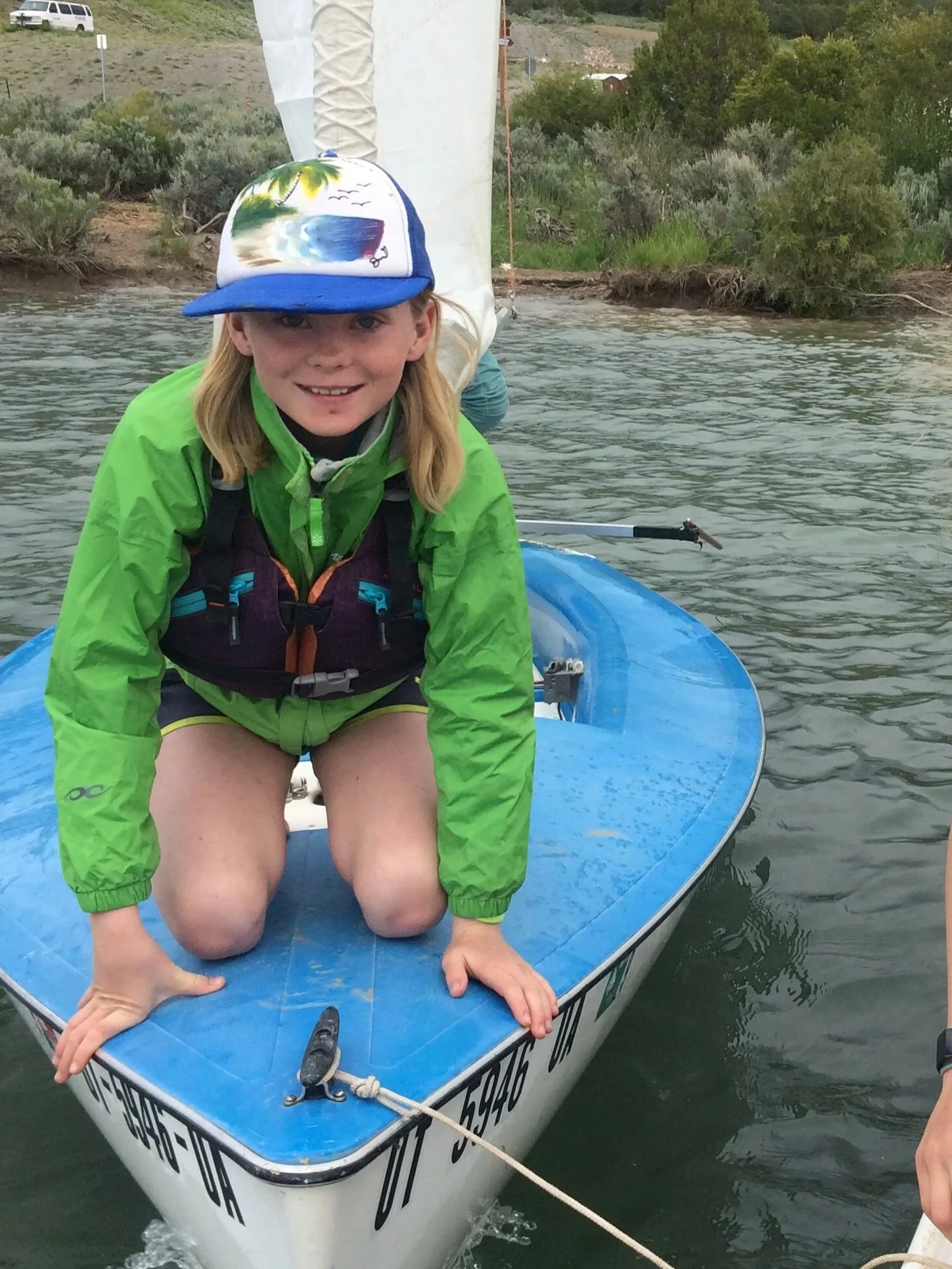 A girl wearing a colorful cap and a bright green jacket on a blue sailboat in a river, with a background of green trees and shrubs.