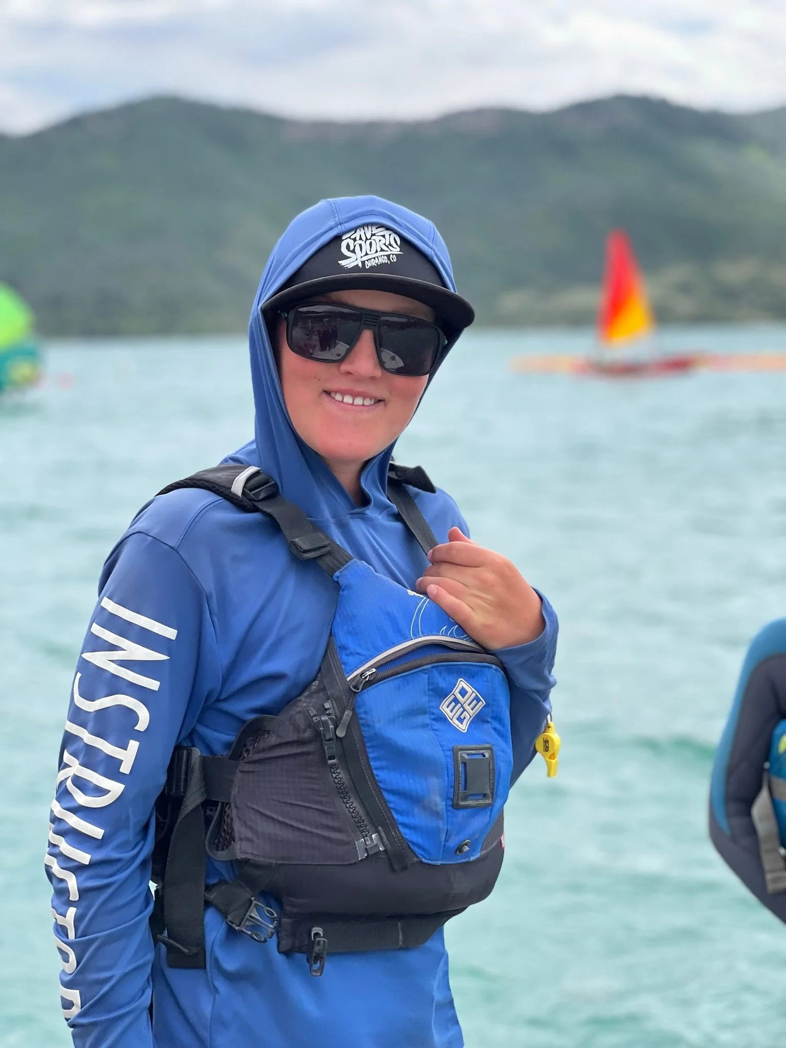 Person in blue outdoor gear with sunglasses and a cap, smiling, on a lake with hills in the background.