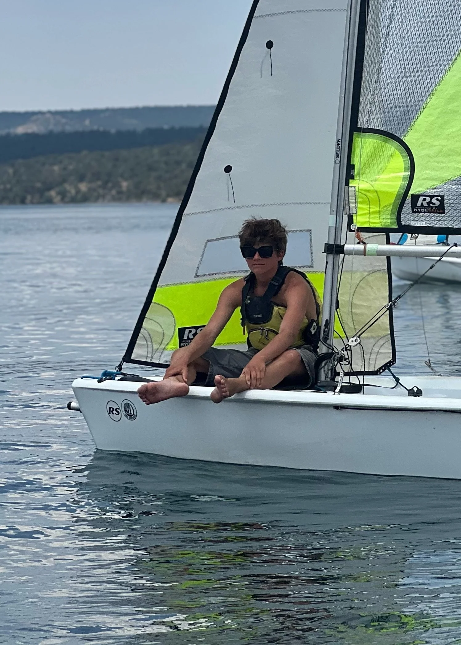A young boy sitting on the bow of a small sailboat on a lake, wearing sunglasses, a life jacket, and shorts, with water and distant hills in the background.