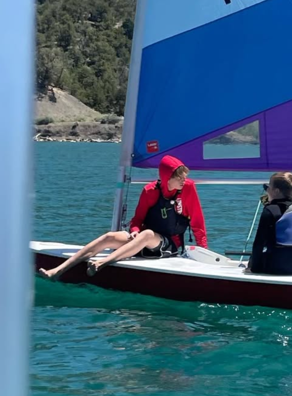 A person sitting on the edge of a small sailboat on a lake, wearing a red hoodie and black shorts, with a blue and purple sail in the background.