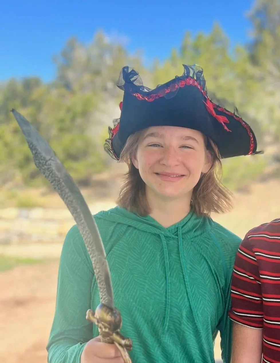 Smiling girl wearing a pirate hat and holding a toy sword outdoors.