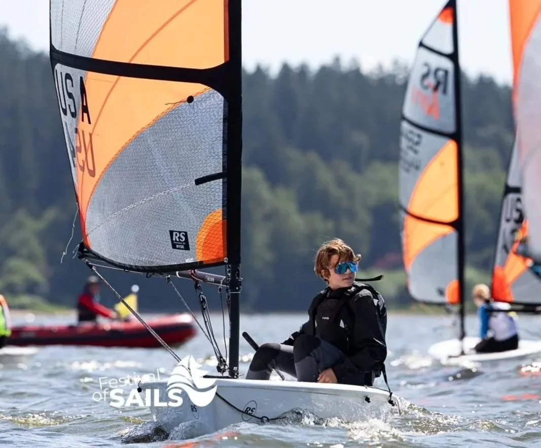 Young person sailing a small sailboat with orange and gray sails on a body of water, with other sailboats and a forested shoreline in the background.