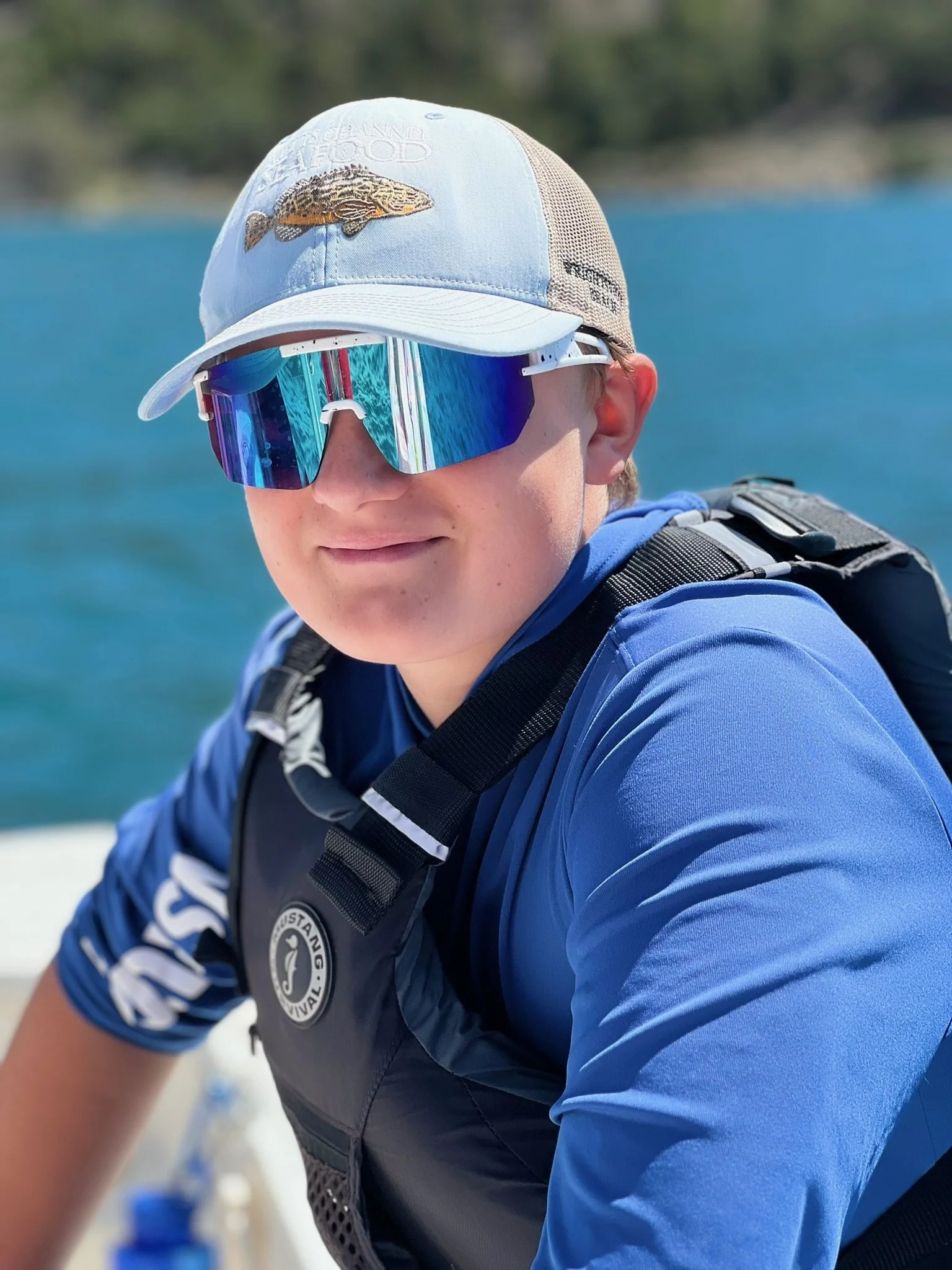 A person wearing a white cap with an embroidered fish and a logo, blue sportswear, reflective sunglasses, and a life jacket, smiling in front of a body of water with a blurred background of trees.