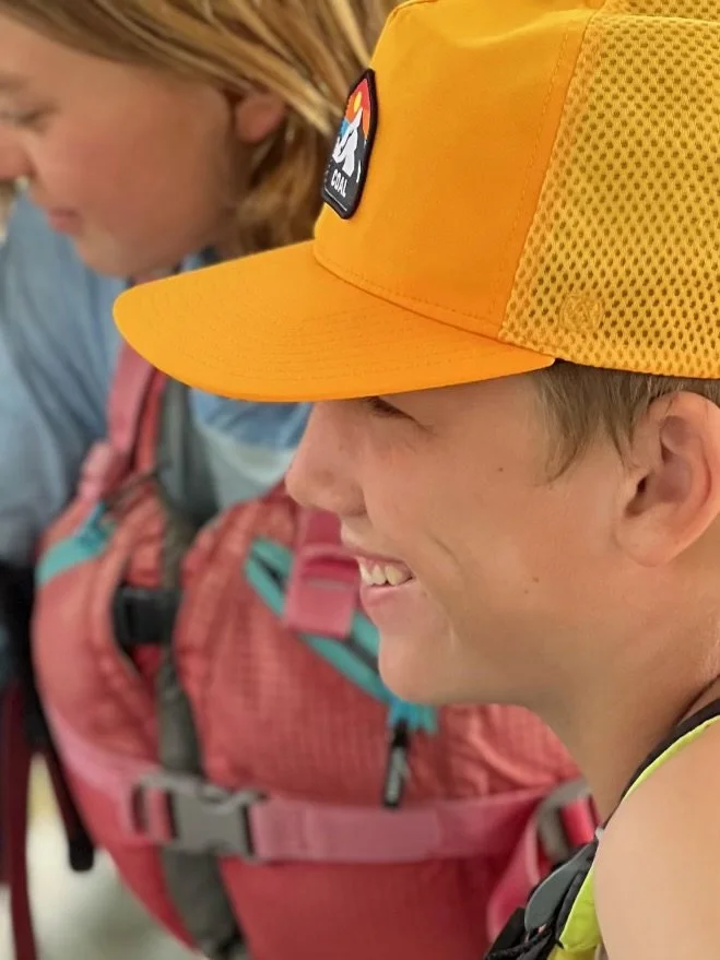 Close-up of a smiling young boy wearing an orange cap with a patch, standing next to a person with a pink backpack.