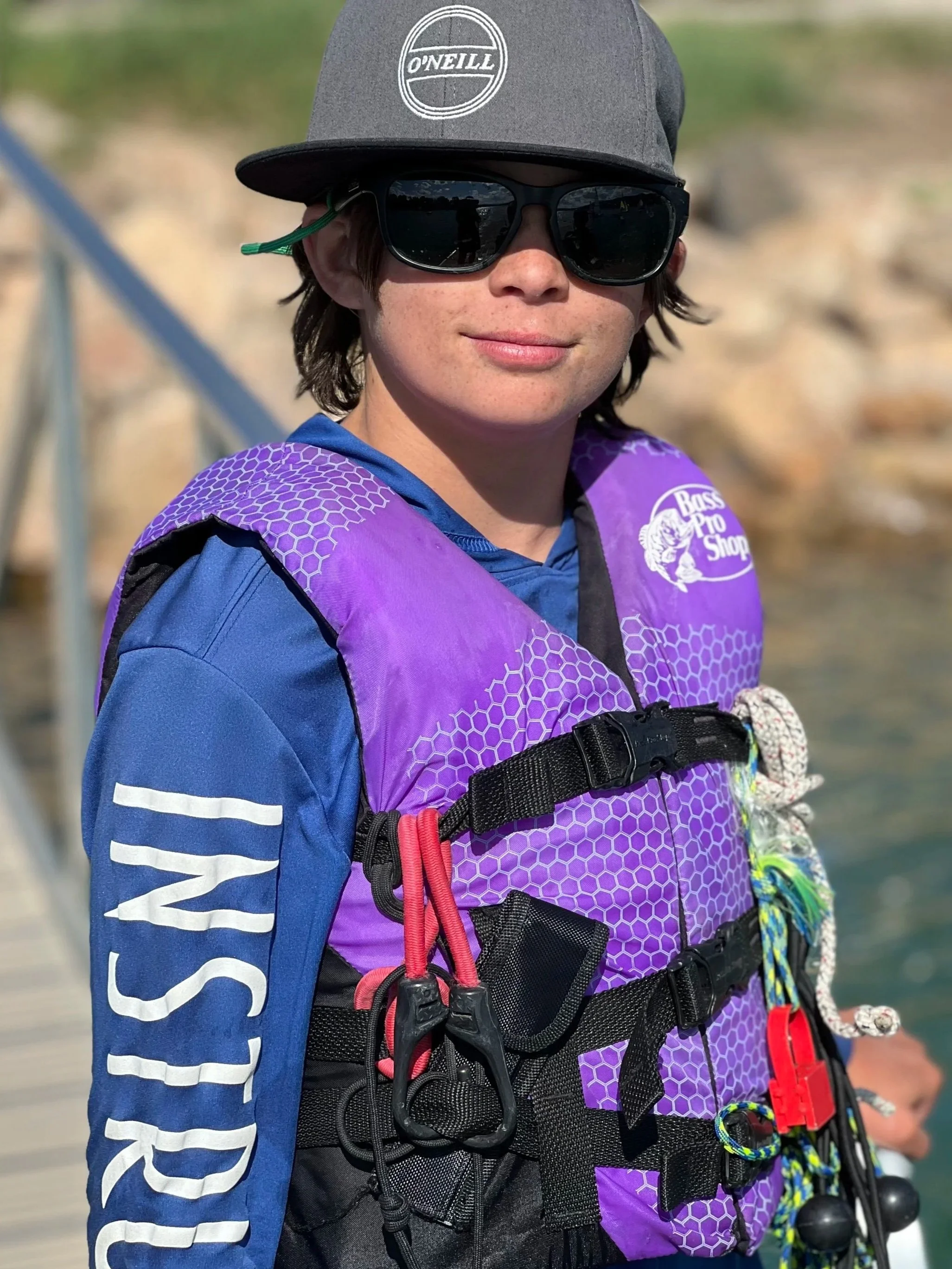 Person wearing sunglasses, a gray cap with 'O'Neill' logo, a blue long-sleeve shirt, and a purple life vest with various straps and chin-up clips. The person is standing outdoors on a dock near water.