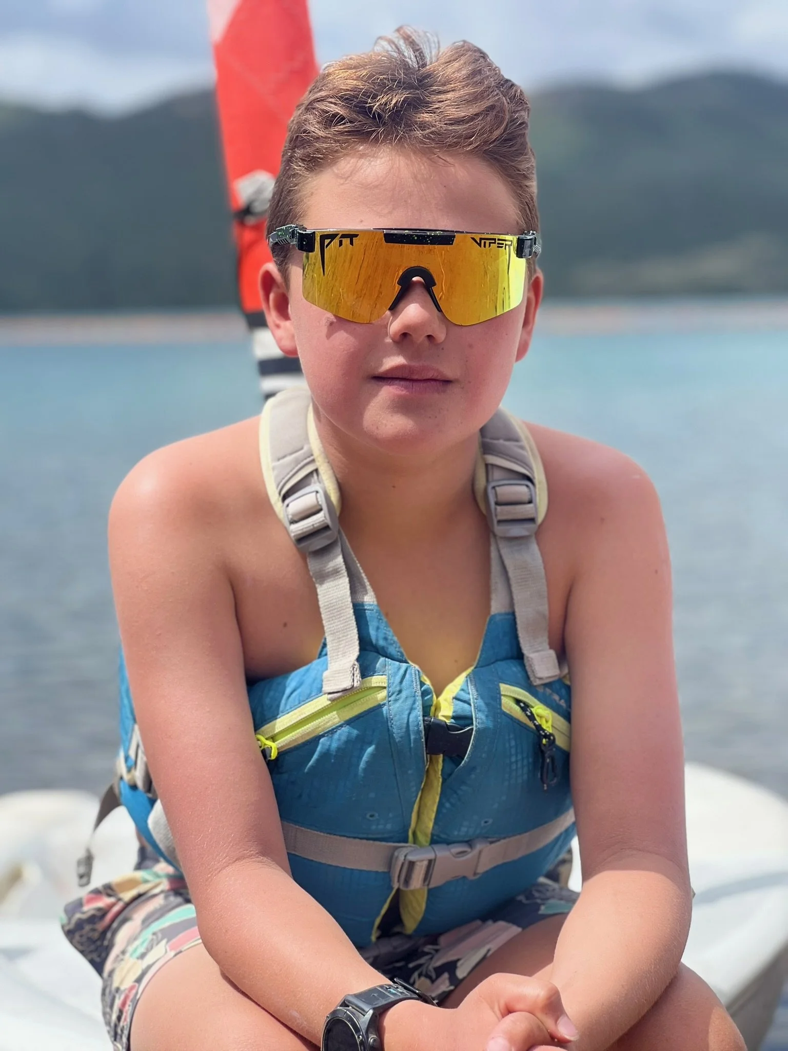 A young boy wearing sunglasses, a life jacket, shorts, and a watch, sitting on a boat near a body of water with hills in the background.