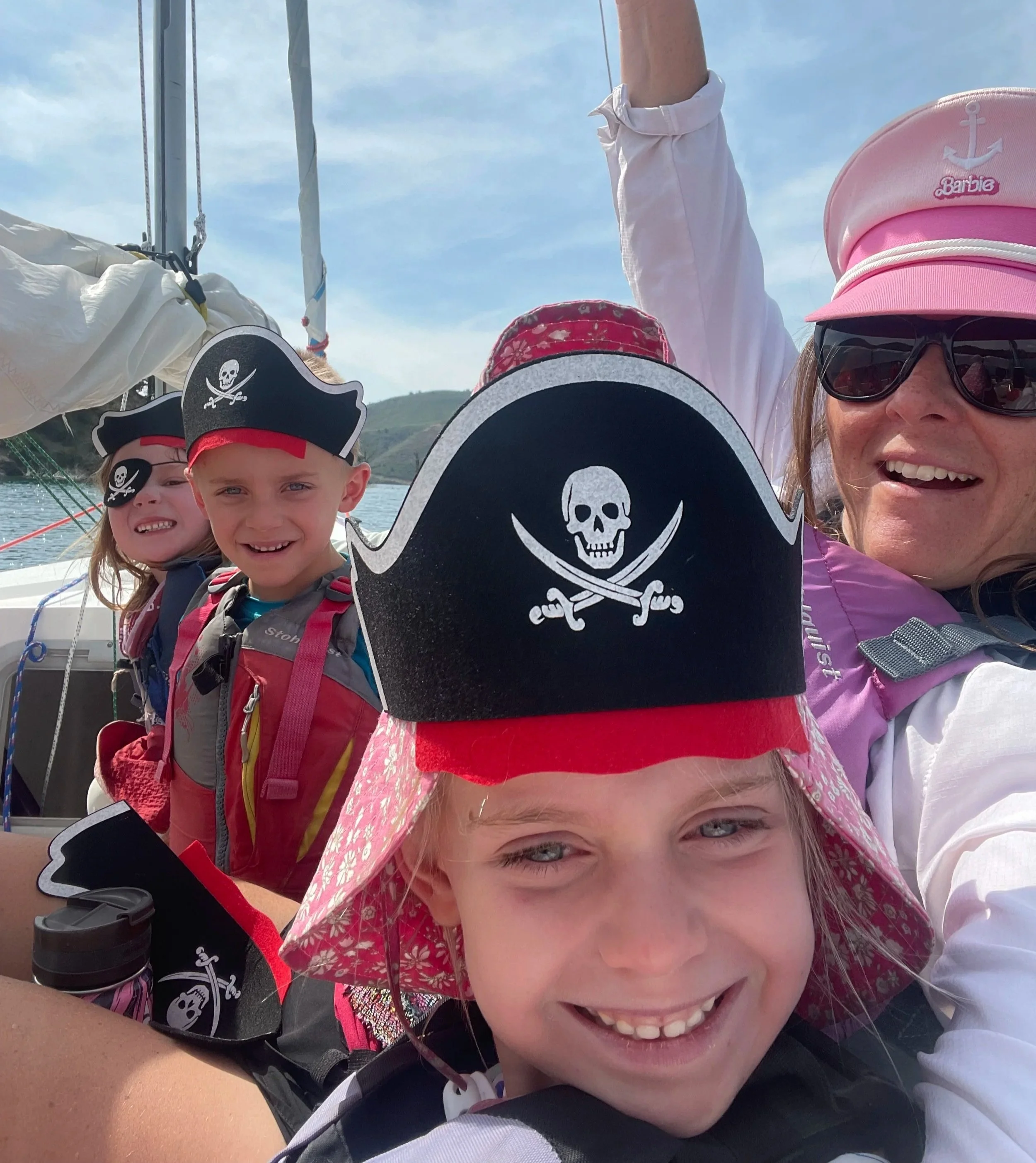 A woman and four children on a boat, all wearing pirate hats with skull and crossbones, enjoying a sunny day on the water with hills in the background.