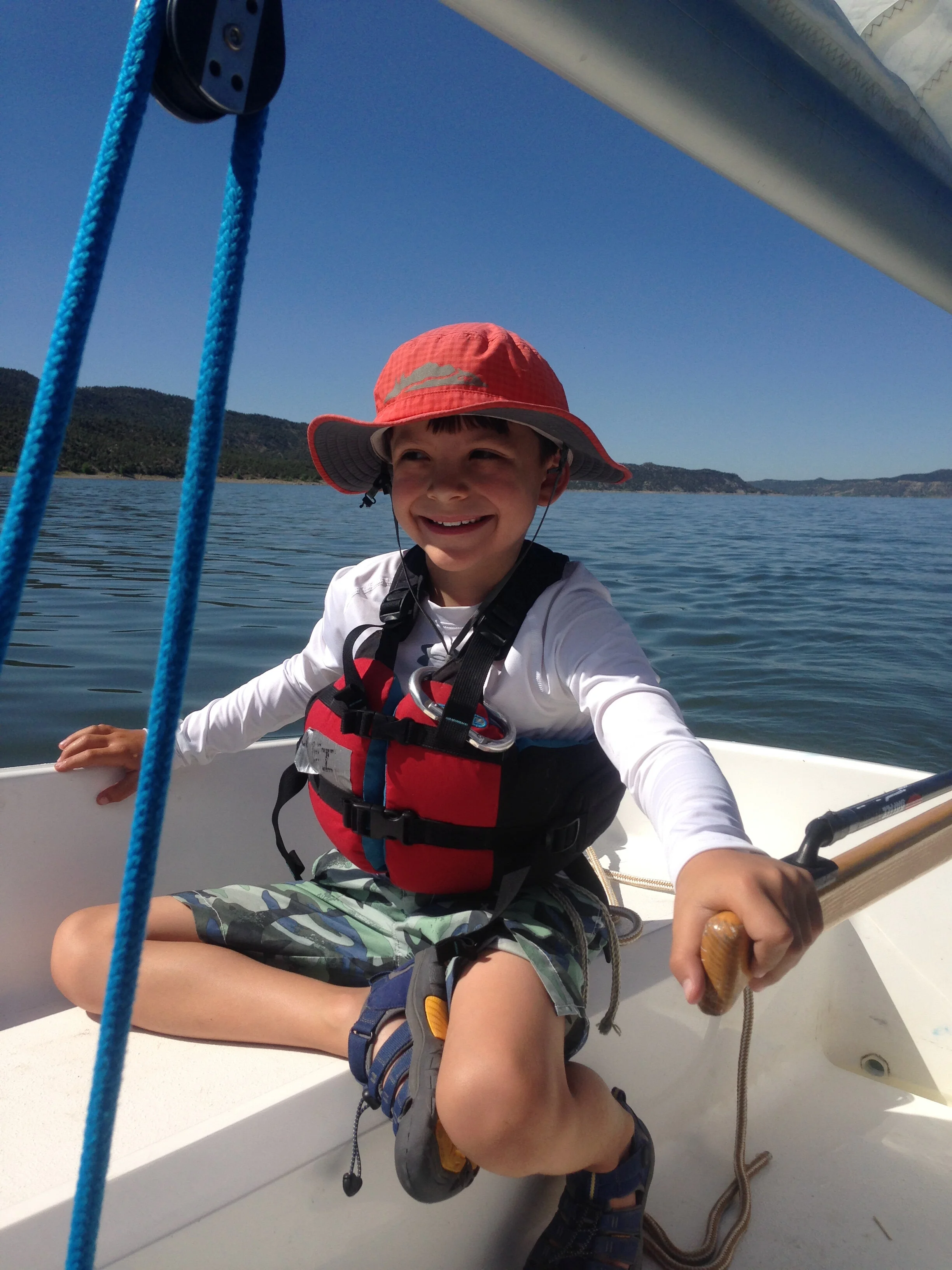 A smiling boy in a red sun hat, life vest, and camo shorts sitting in a small boat on the water during summer camp.