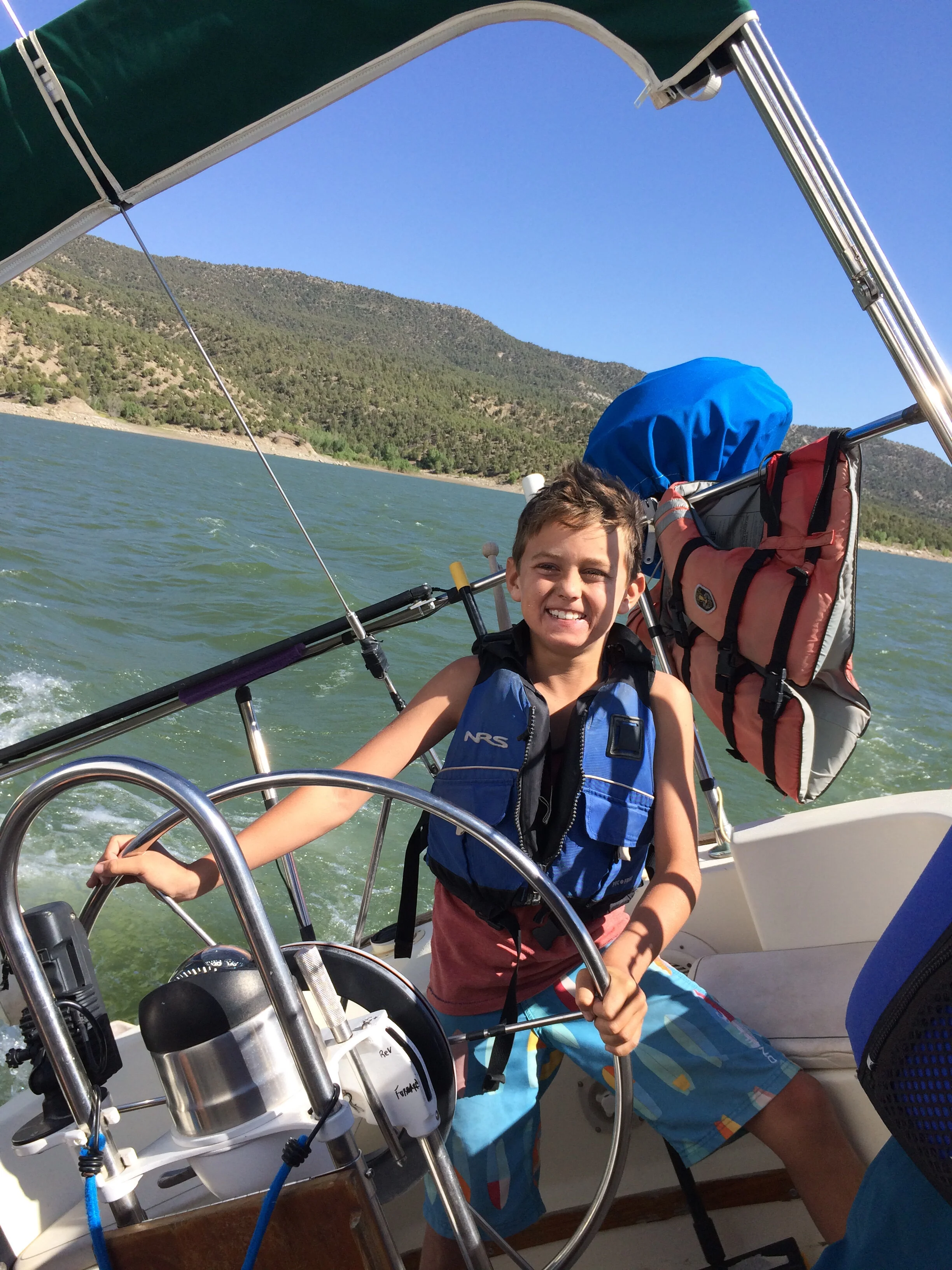 Smiling young boy holding steering wheel on a boat, wearing a blue life jacket and colorful swim shorts, with a mountainous landscape in the background under a clear blue sky, while at summer camp.
