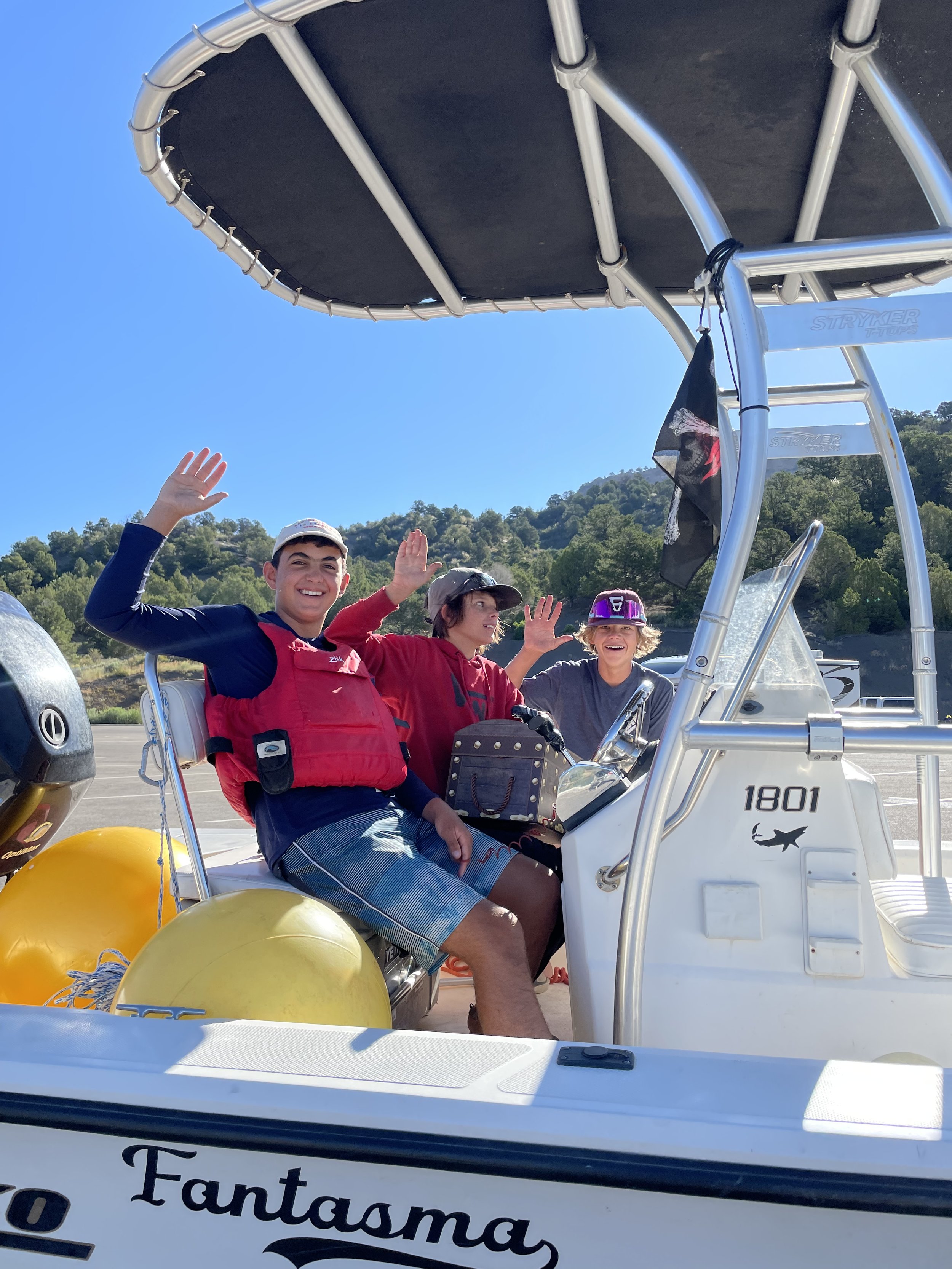 Three teenagers on a boat, smiling and waving, with a mountainous background and clear blue sky.