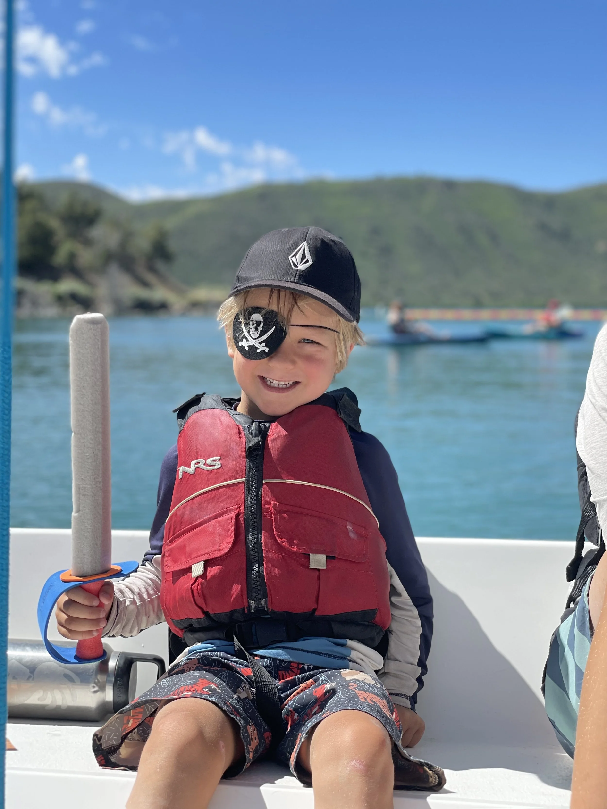 A young boy wearing a pirate eye patch, a black cap with a logo, a red life jacket, and colorful shorts, sitting on a boat holding a foam sword, smiling at the camera with a scenic water and hillside background.