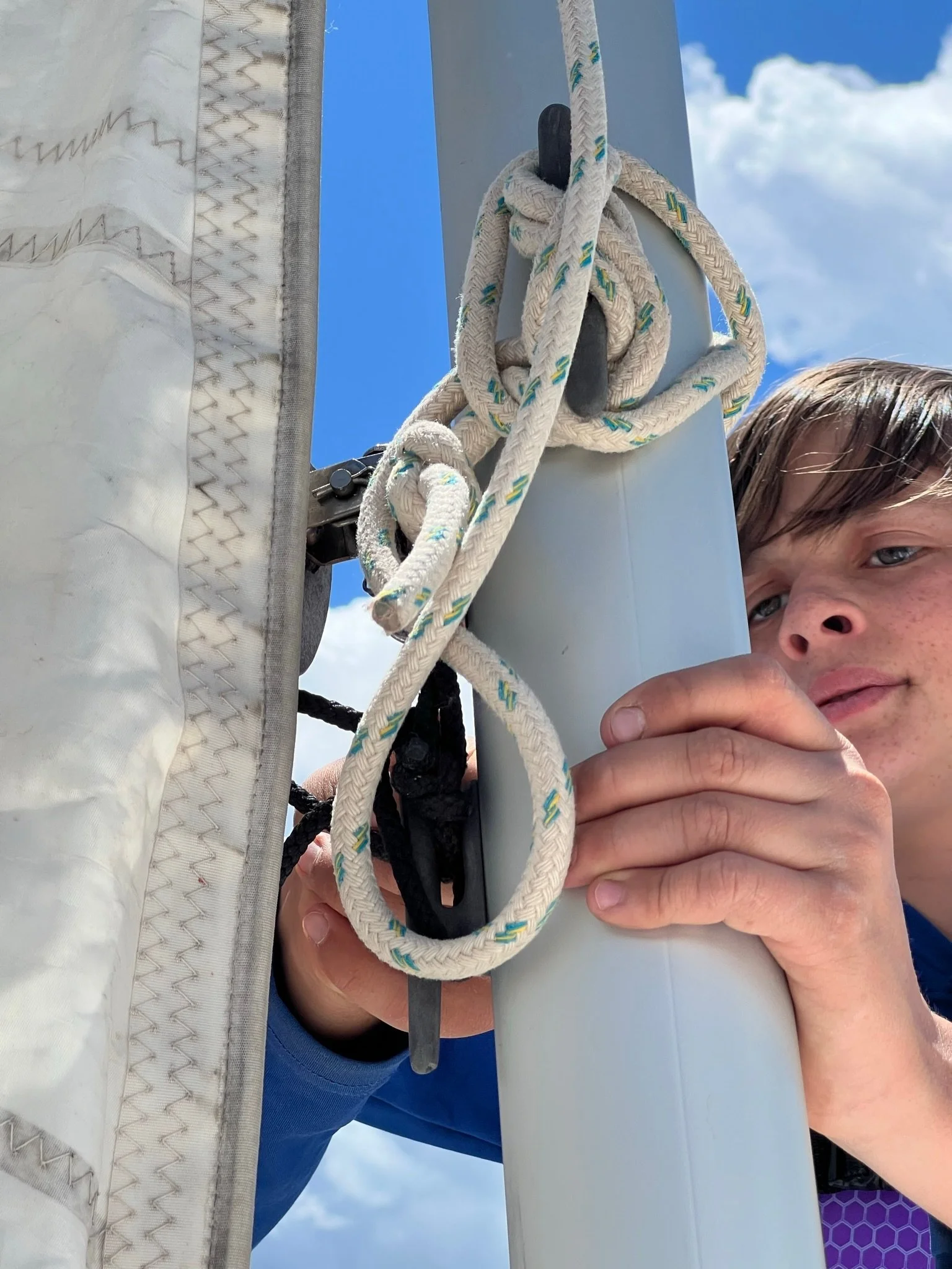 A person with short brown hair and blue eyes attaching a rope to a mast on a sailboat under a partly cloudy sky.