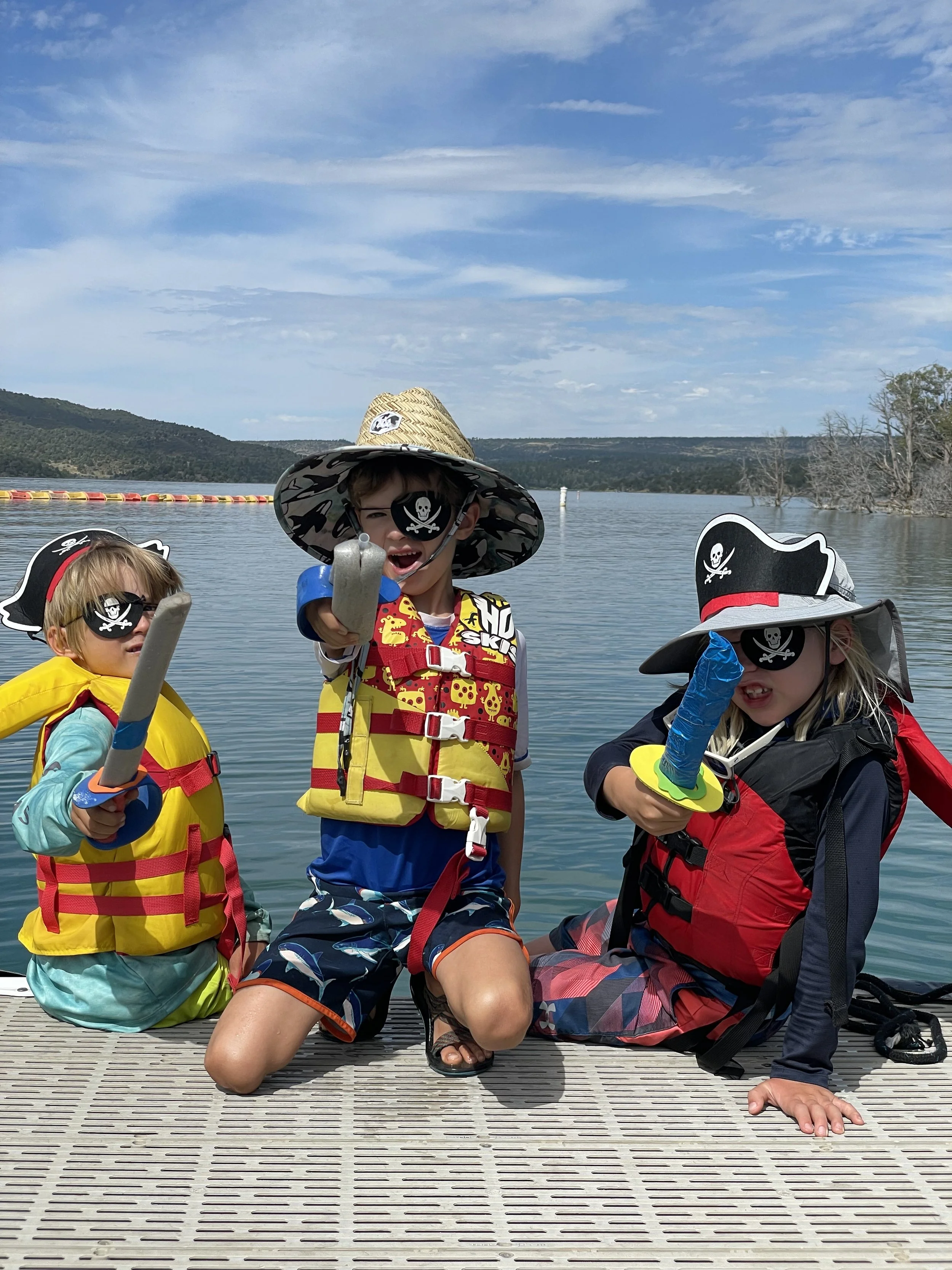 Three children dressed as pirates on a dock by a lake, wearing pirate hats with skull and crossbones, holding foam swords, with water, mountains, and a partly cloudy sky in the background.