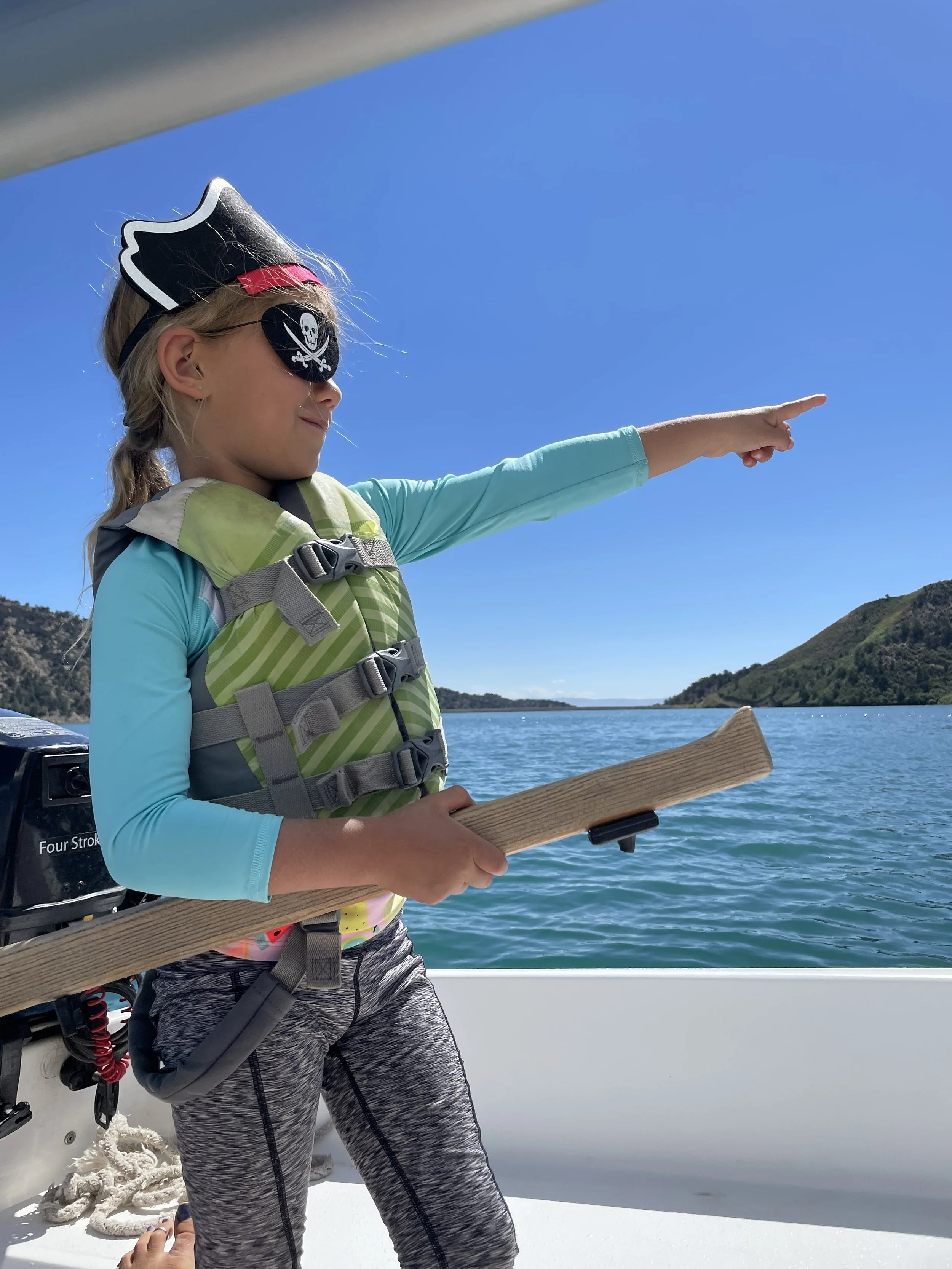 A young girl wearing a pirate eye patch, a pirate hat, and a life jacket is on a boat, pointing toward the water with a paddle in hand, overlooking a lake or bay with hills in the background.