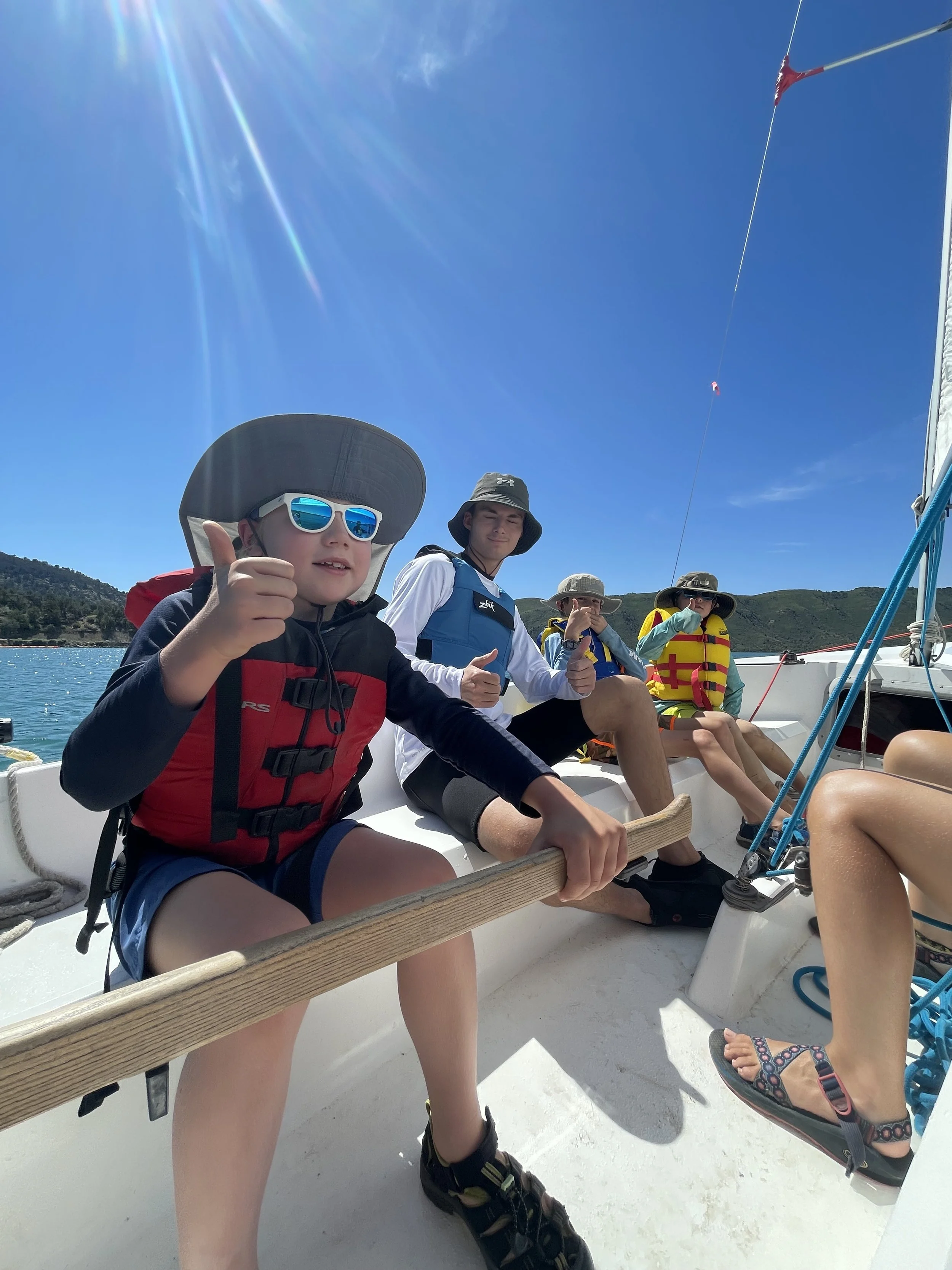 Group of children on a boat wearing life jackets and hats, enjoying a sunny day outdoors.