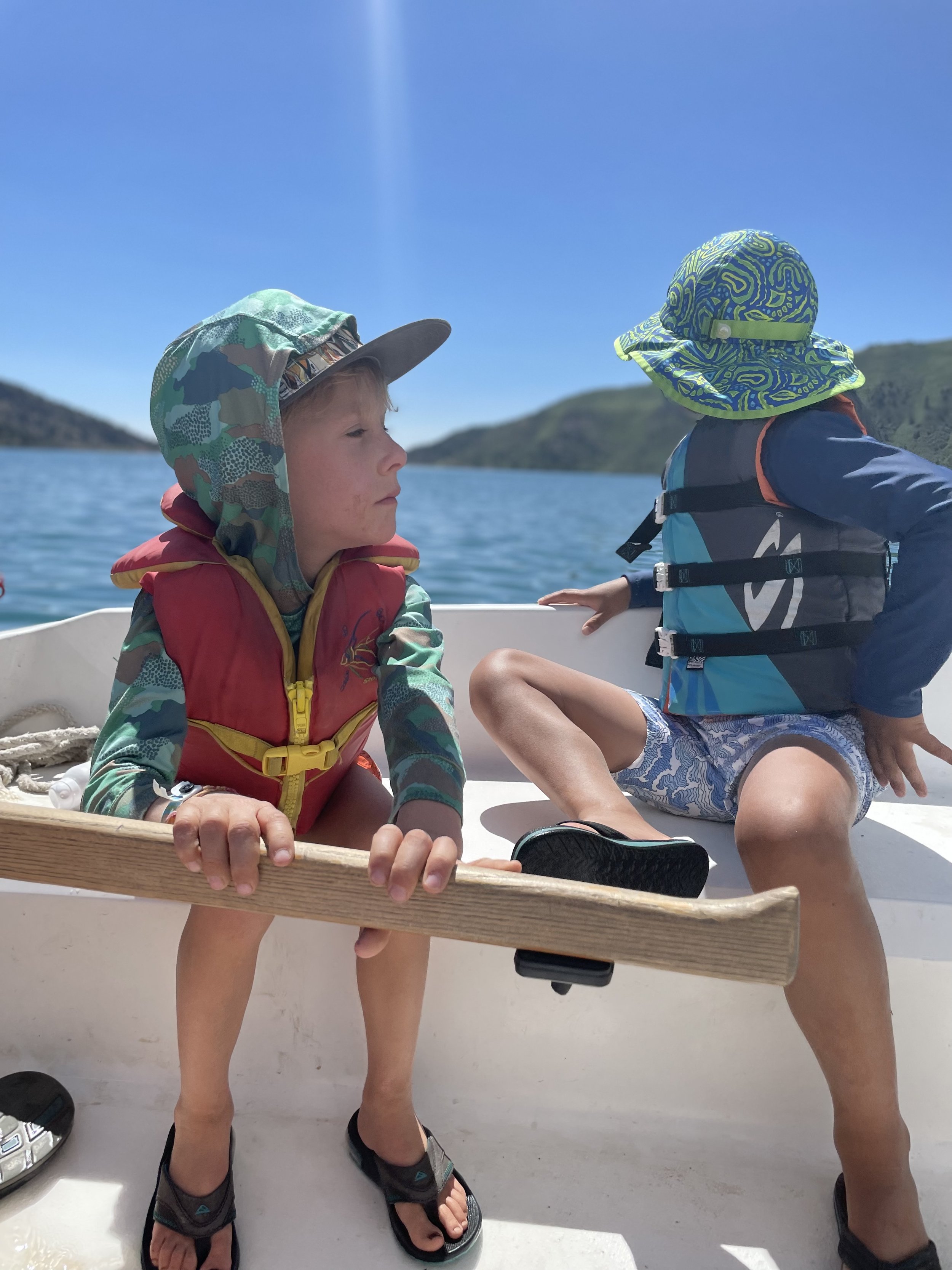 Two young children sitting on a boat, wearing sun hats, life vests, and sandals, with a lake and mountains in the background on a sunny day.