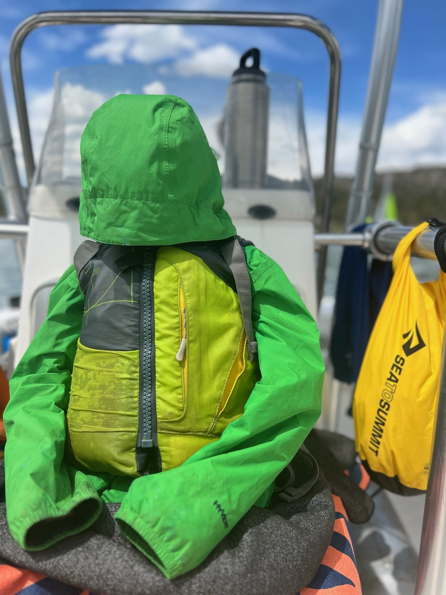 A green waterproof jacket with a hood, a yellow and gray backpack, and a yellow dry bag hanging on a boat with a metal railing, sky with clouds in the background.