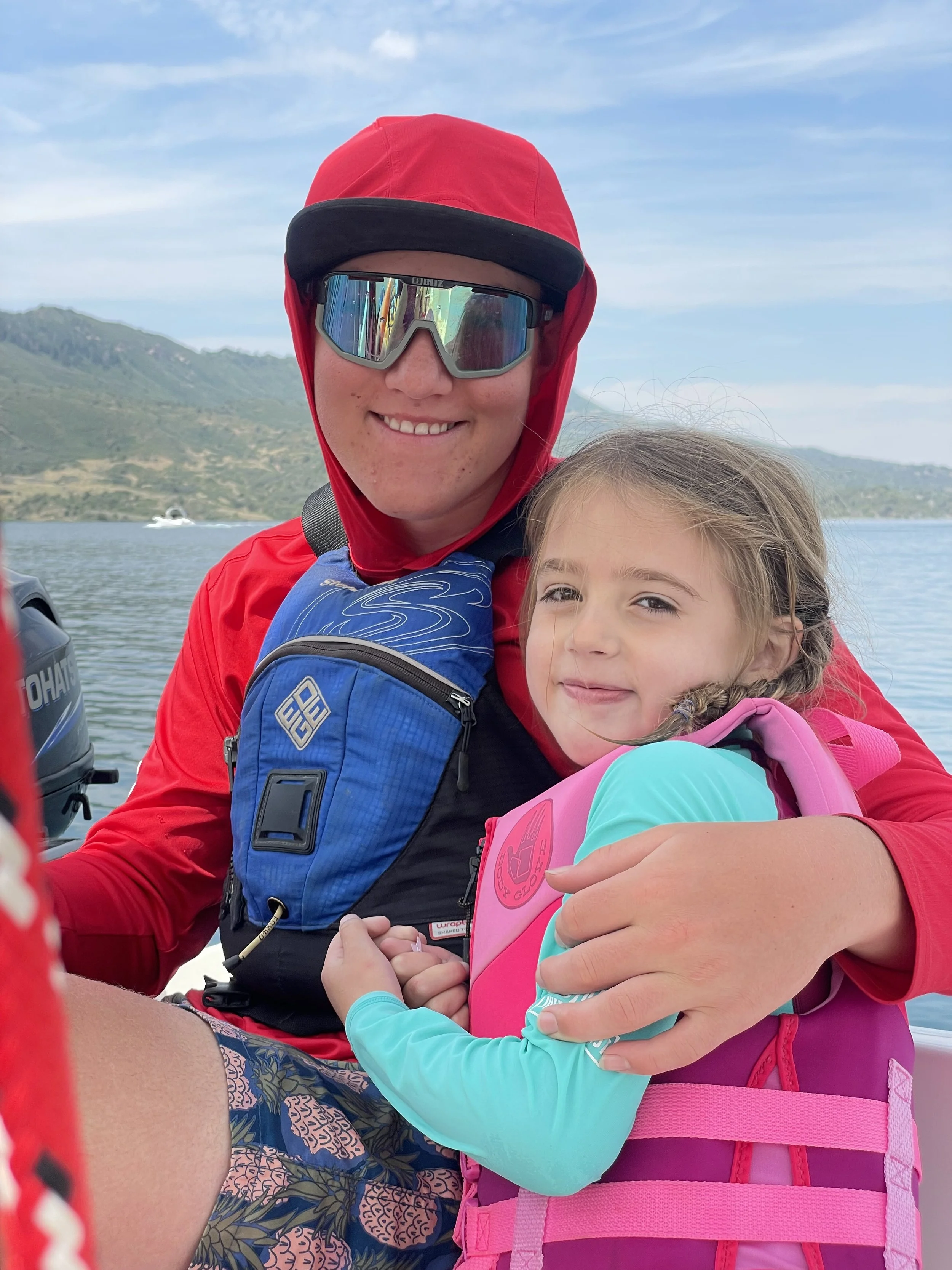 A woman and a young girl on a boat, with the woman wearing sunglasses and a red hooded jacket, smiling at the camera. The girl is wearing a pink life jacket and a blue long-sleeve shirt, clinging to the woman, with a lake and hills in the background.