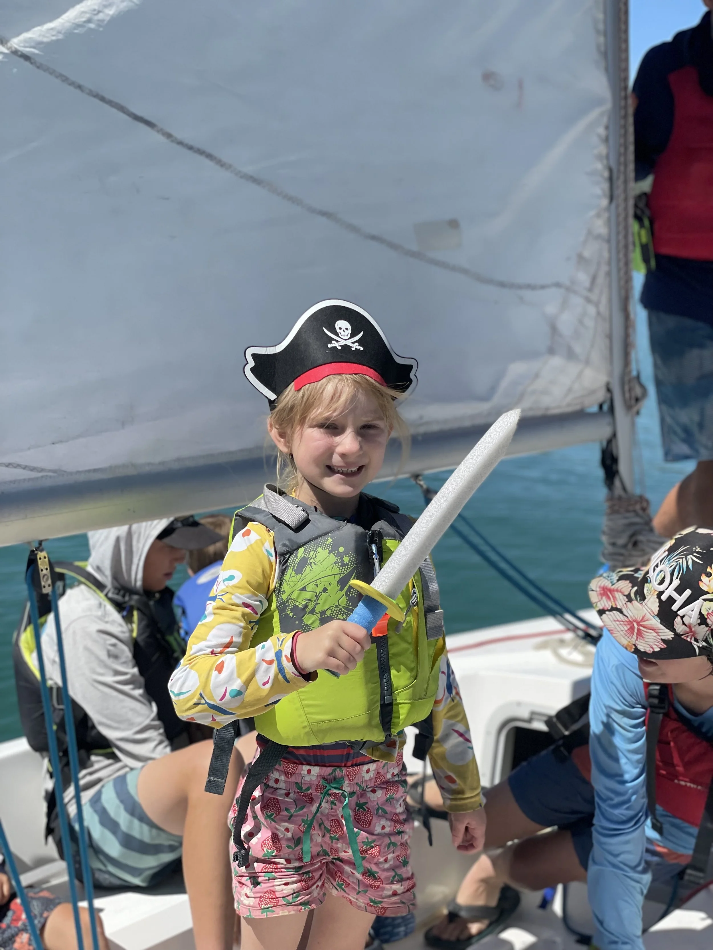 A young girl dressed as a pirate with a skull and crossbones hat, holding a sword, on a boat with other children.