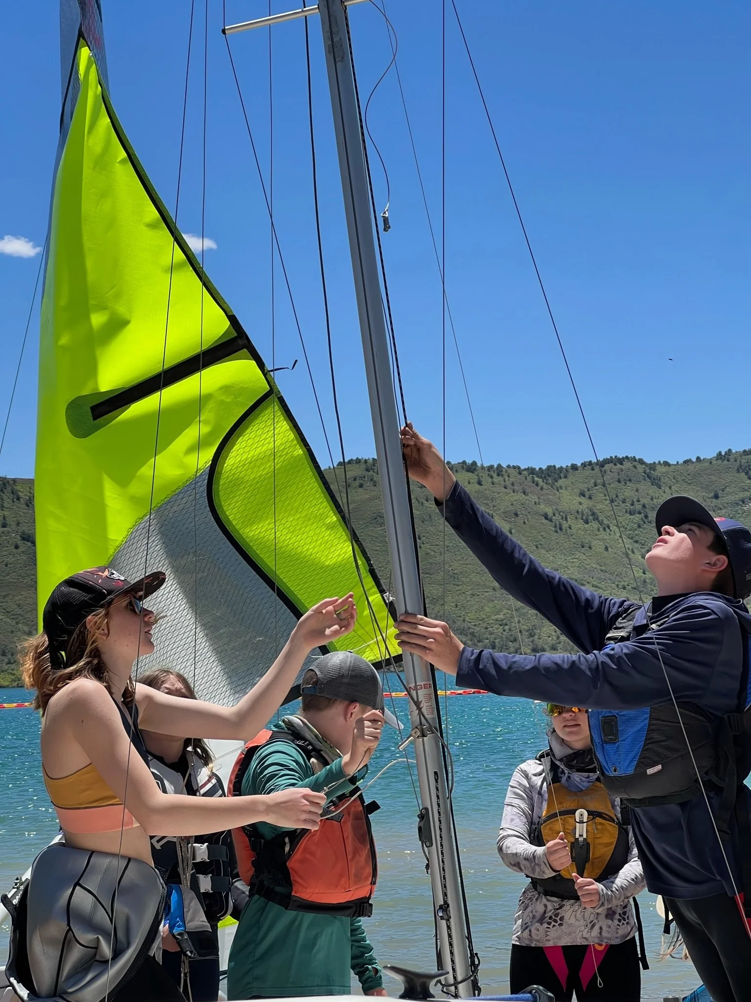 Group of kids preparing a sailboat near a lake with mountains in the background on a sunny day.