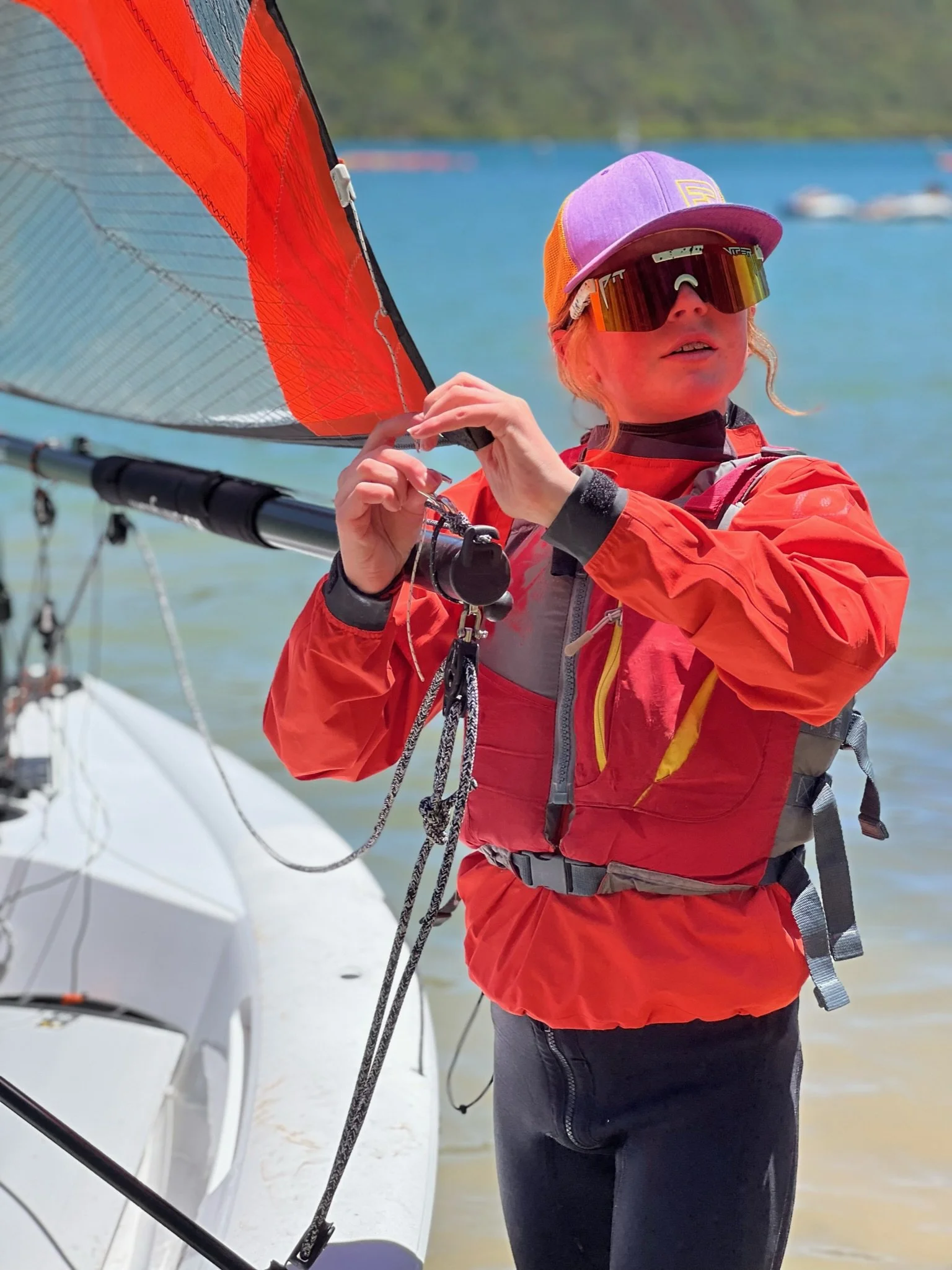 A woman on a boat wearing sunglasses, a purple cap, and a red jacket, adjusting sailing equipment on a sailboat with water and other boats in the background.