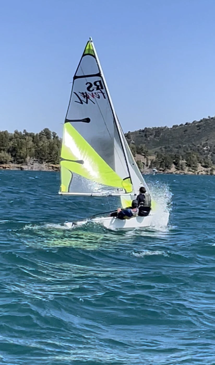 Two people sailing a small sailboat with bright yellow and white sails on clear blue water, with a hilly shoreline and trees in the background under a cloudless sky.