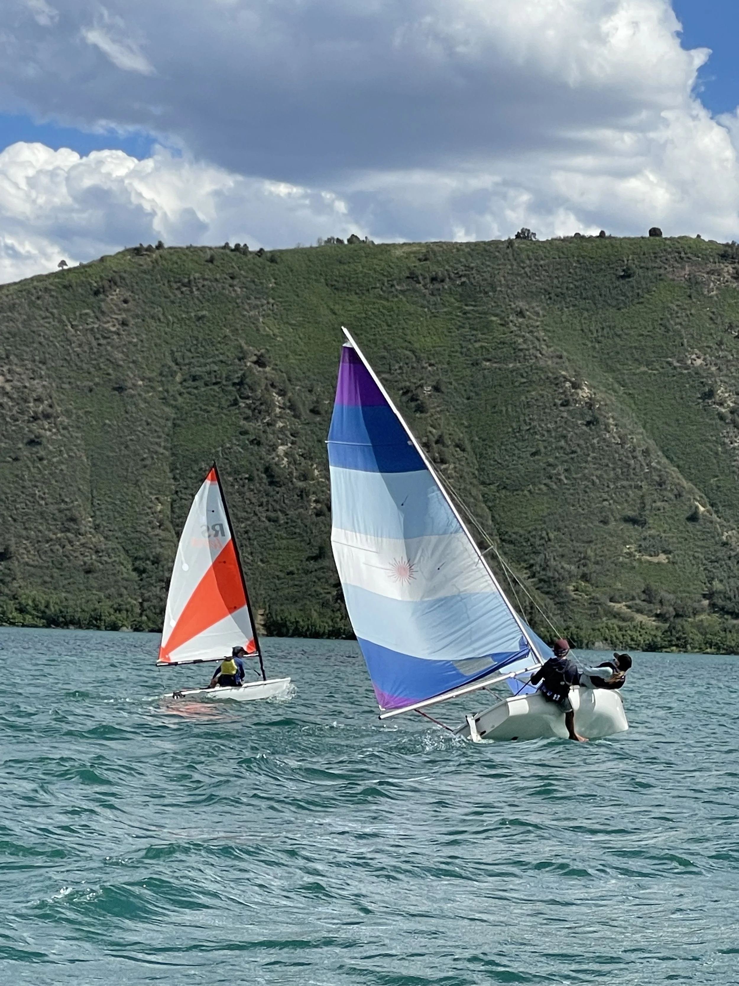 Two people sailing on a large body of water with green hills and a partly cloudy sky in the background.