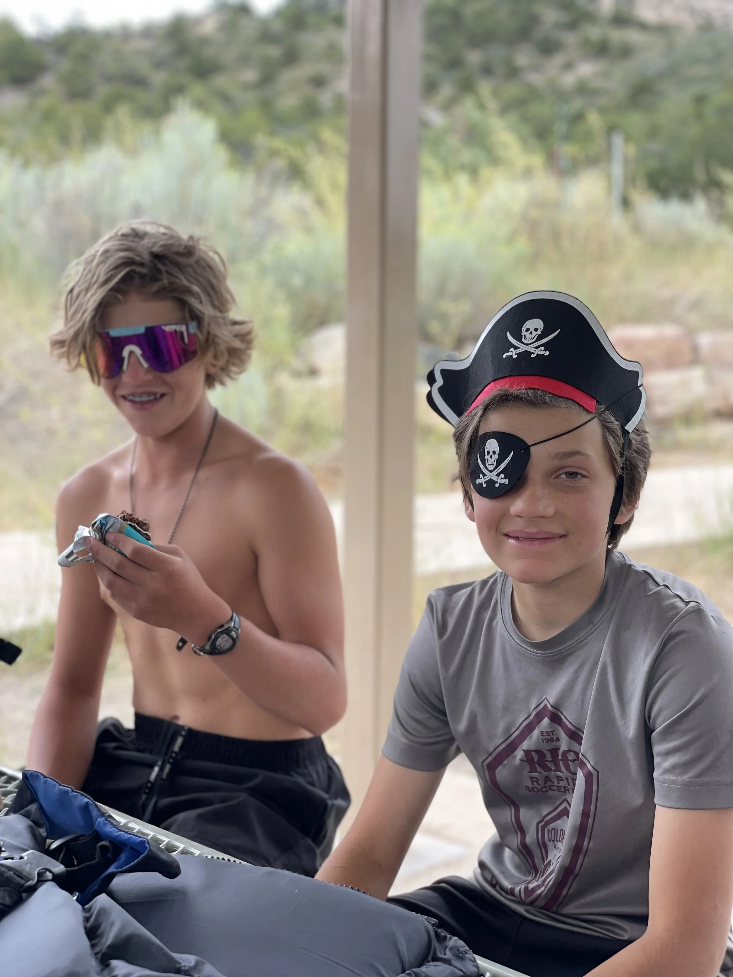 Two boys dressed as pirates, with pirate hats and eye patch, sitting outdoors at a table with a backpack, smiling and enjoying their time.