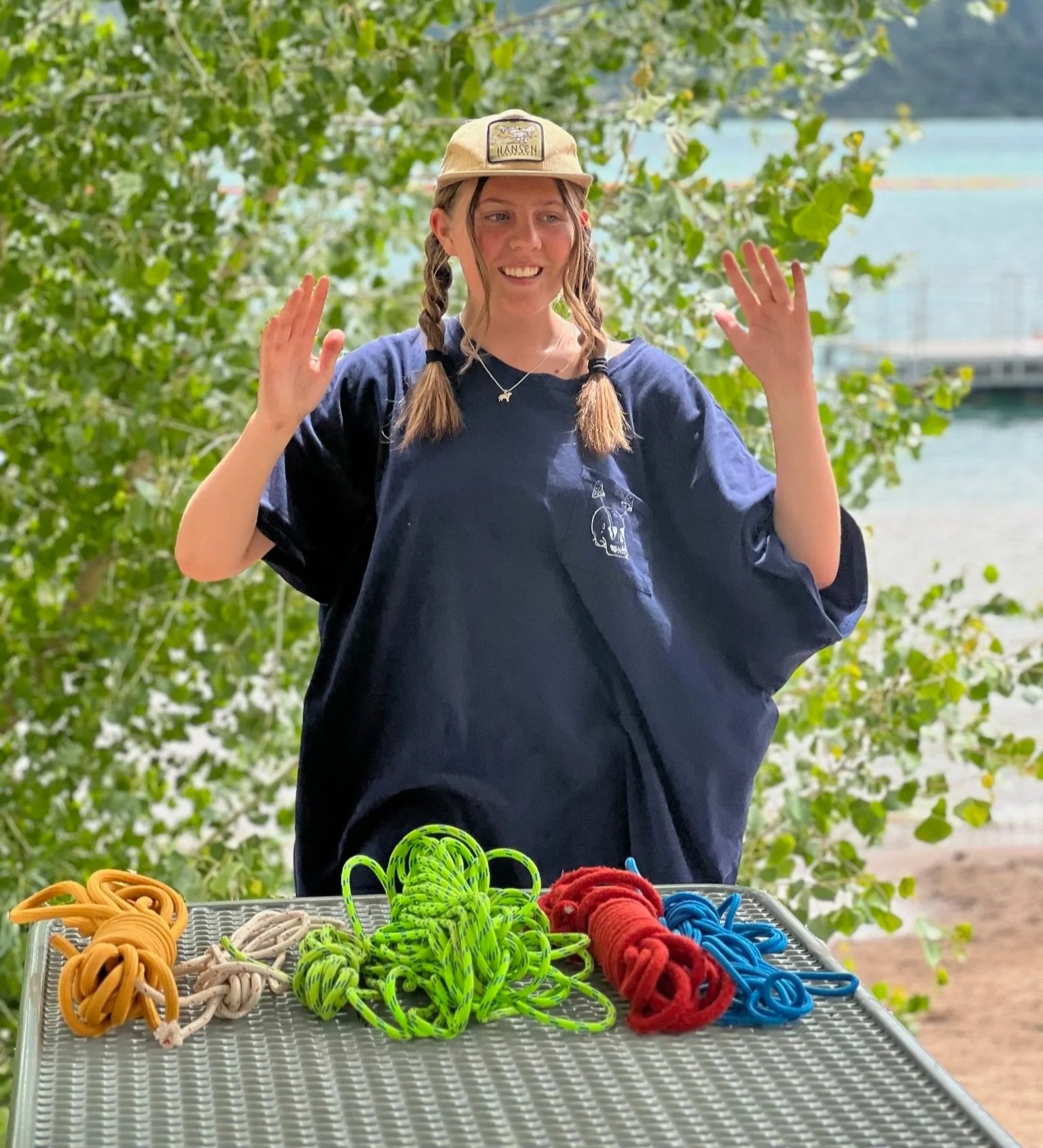 A young woman with pigtails, wearing a blue t-shirt and a tan hat, stands outdoors near water with a tree behind her. She is smiling with her hands raised. In front of her on a table are various colorful ropes, including orange, white, green, red, an
