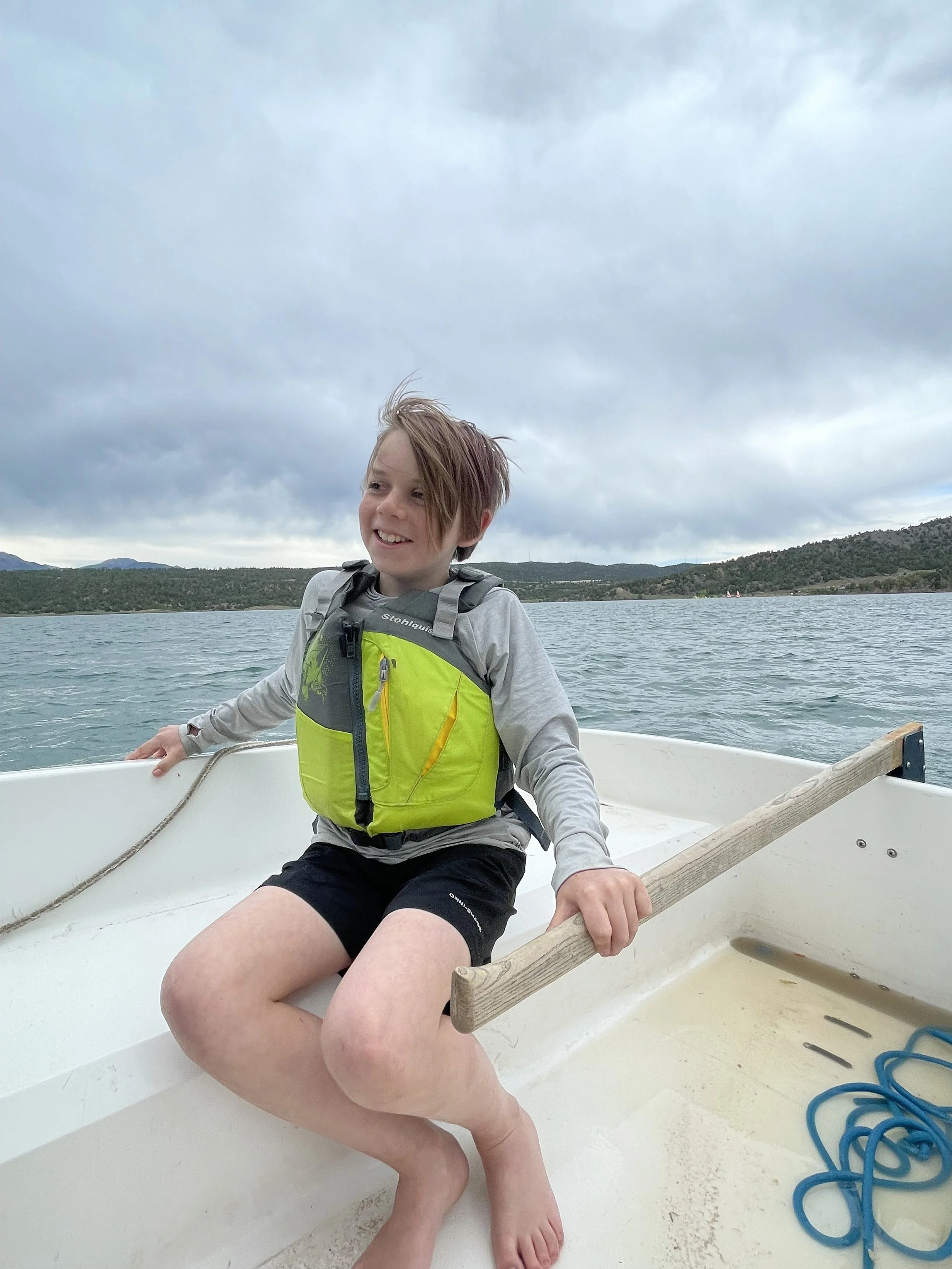 A young boy sitting in a small boat on a lake, holding an oar, with cloudy skies and distant hills in the background.