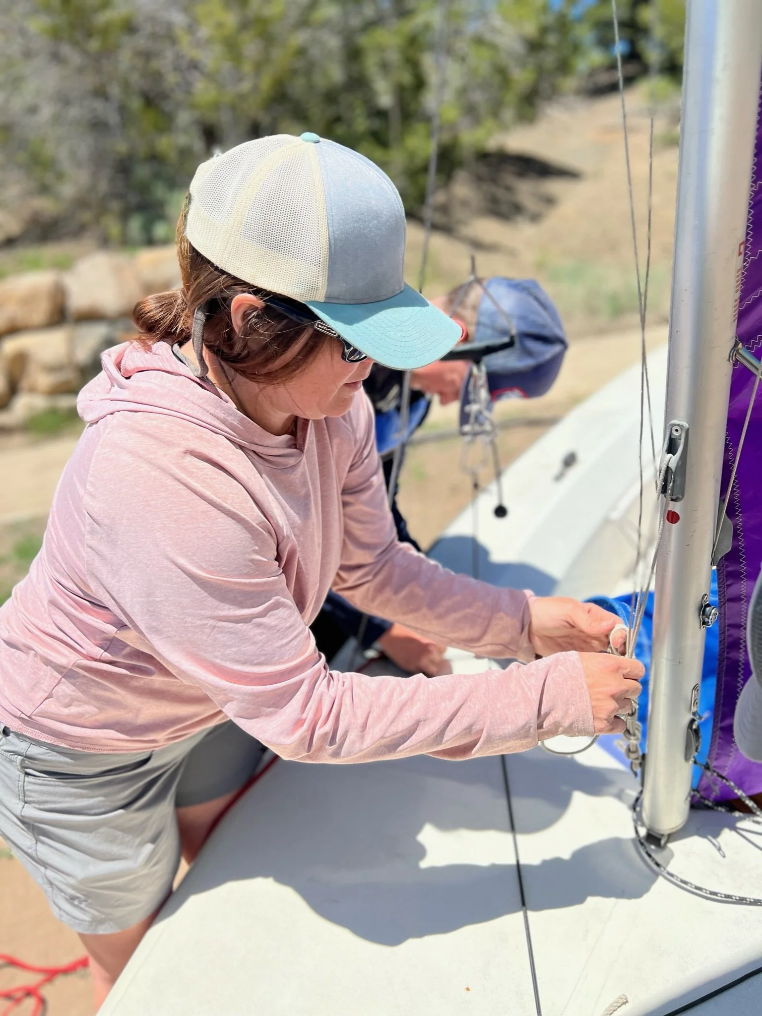 A woman wearing a gray and teal cap, sunglasses, a pink hoodie, and shorts, adjusting the rigging of a sailboat on a sunny day. There is another person in the background, partially visible, also working on the sailboat.