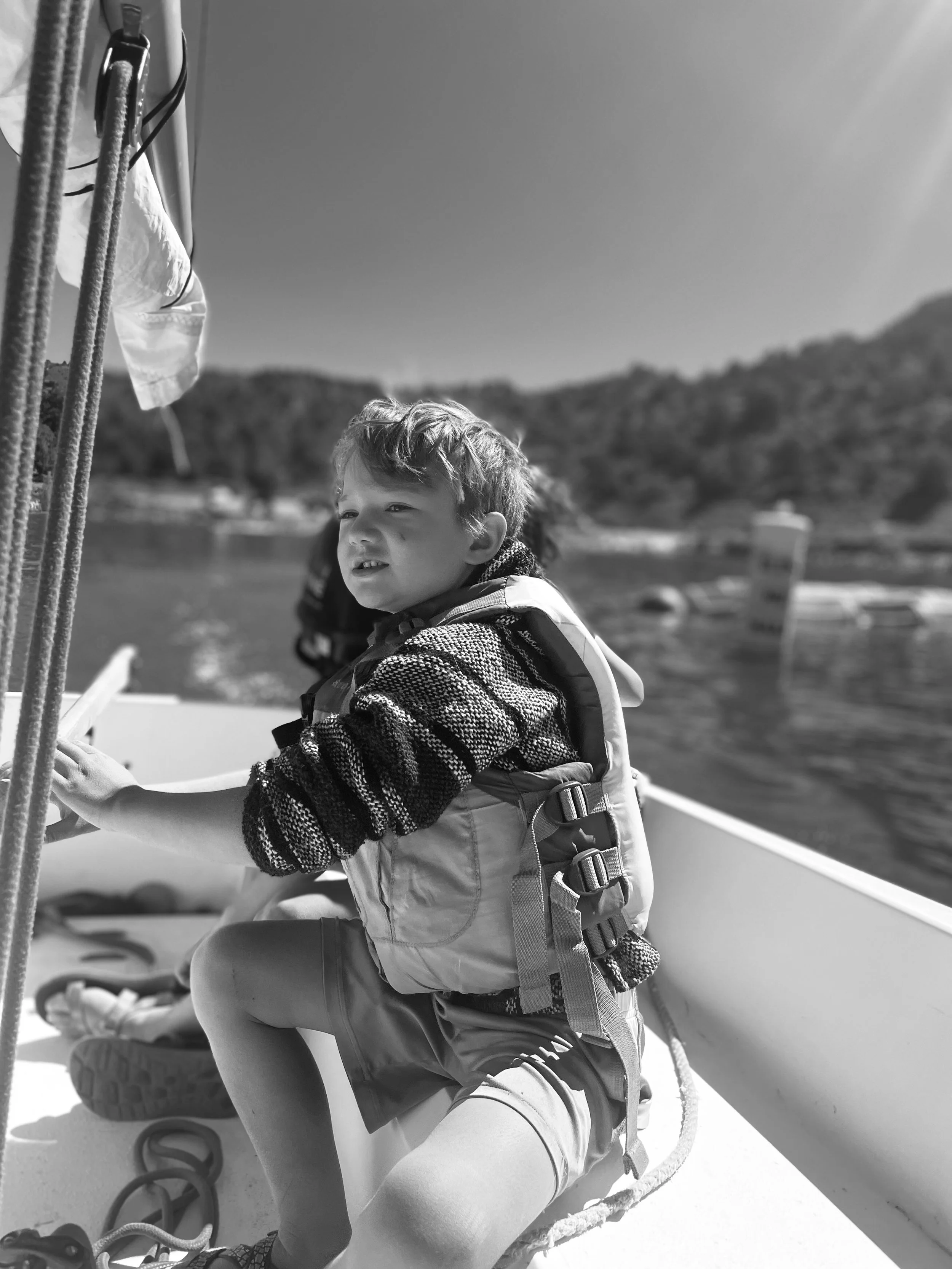 A young boy sitting on a boat, looking out at the water and landscape, wearing a life jacket and casual clothing, with ropes and boat equipment nearby.