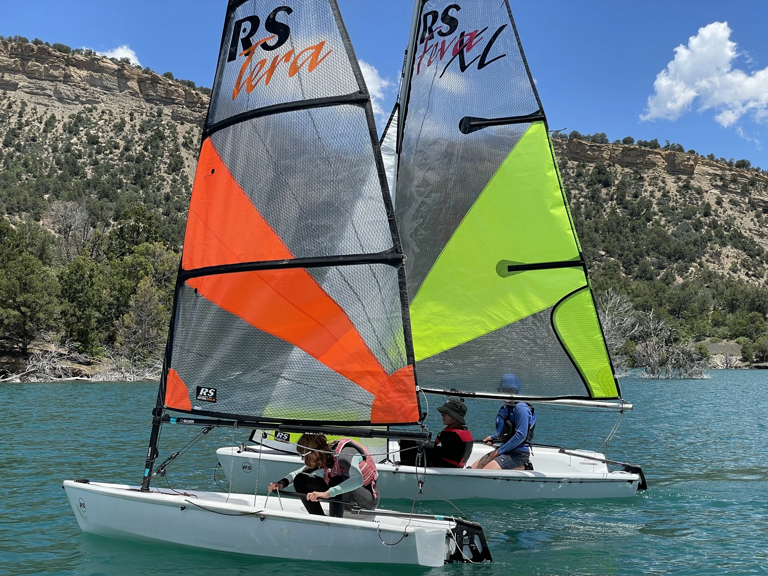 Two small sailboats with colorful sails on a lake, with a rocky hillside and trees in the background.