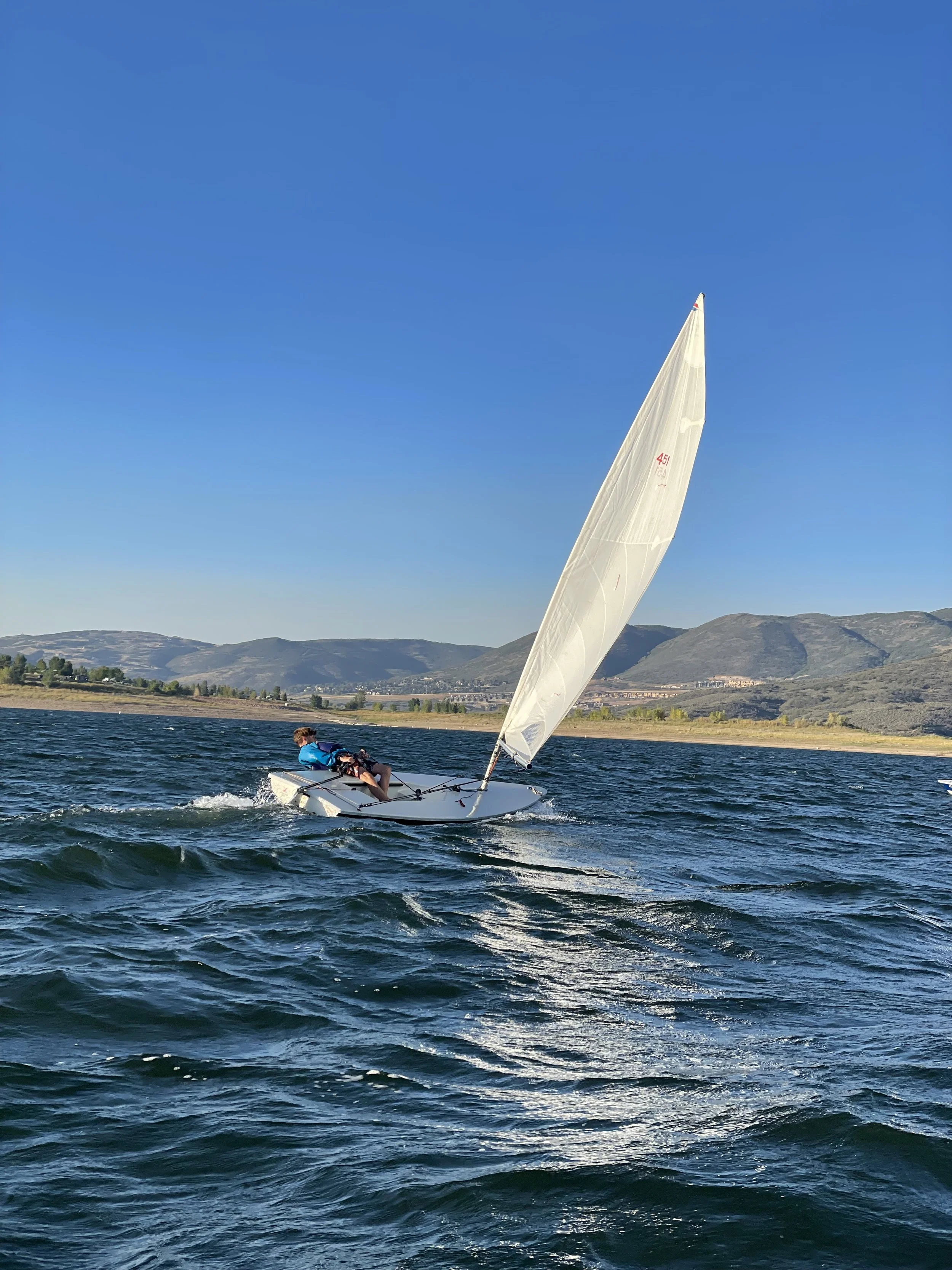 A person sailing a small sailboat on a body of water with hills in the background under a clear blue sky.