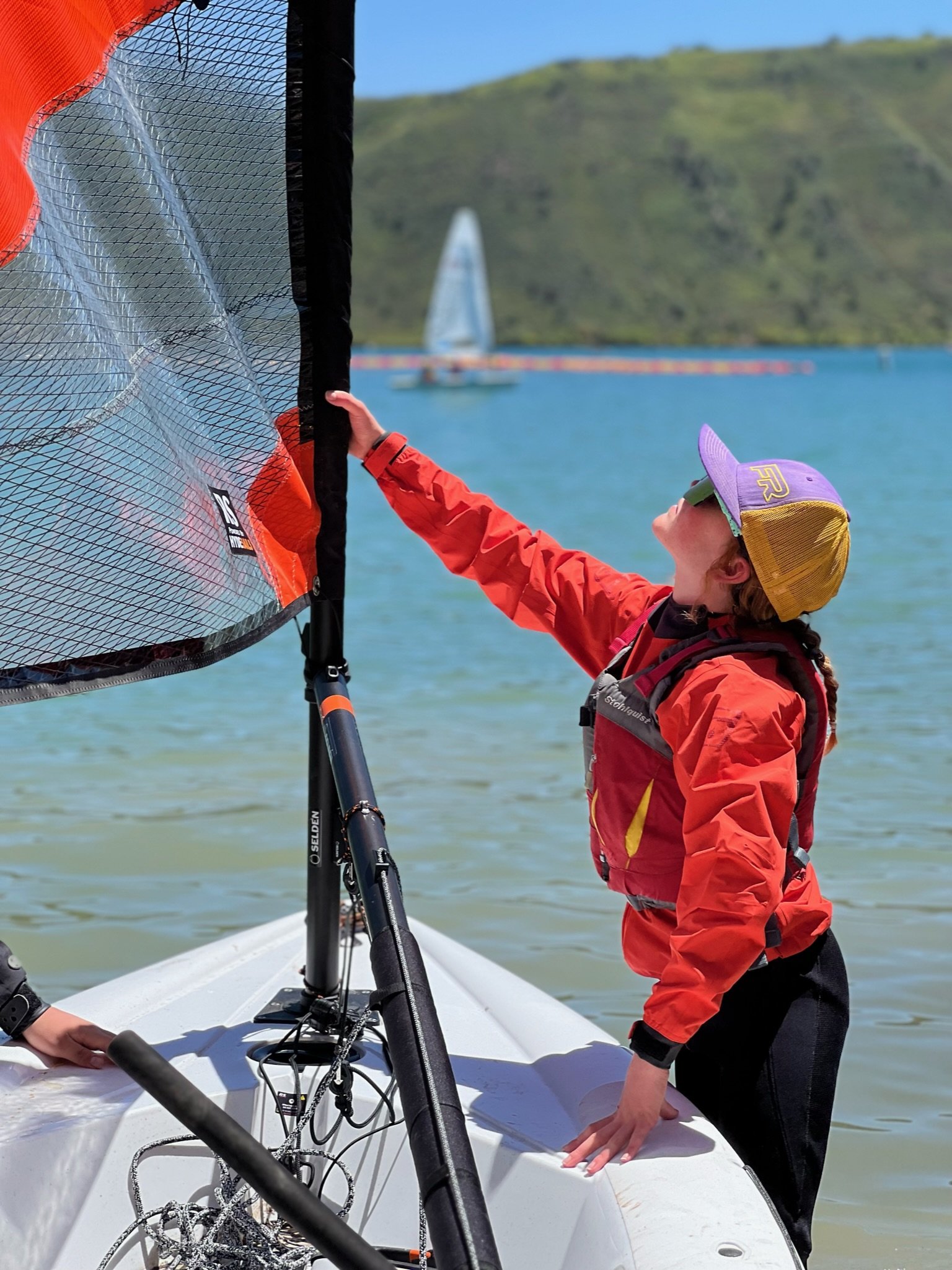 A person in a red and black life jacket and sunglasses adjusting the sail on a small sailboat near a body of water, with a sailboat in the background and green hills beyond.