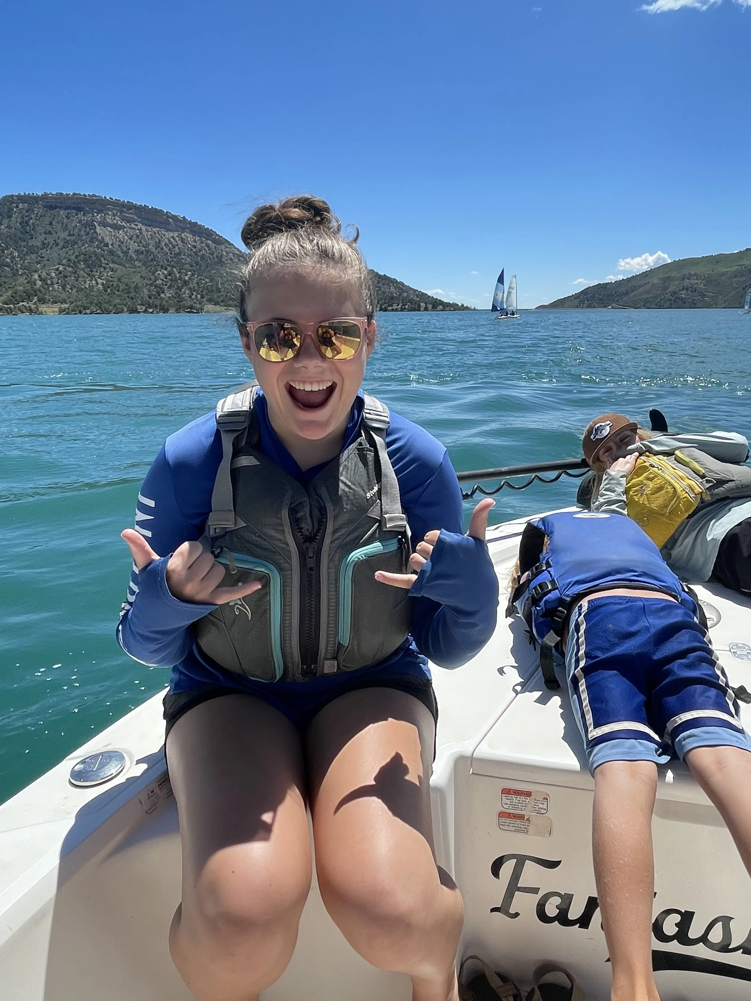 A young woman smiling and making a shaka sign while sitting on a boat with kids in swimsuits, surrounded by blue water and hills, under a clear sky.