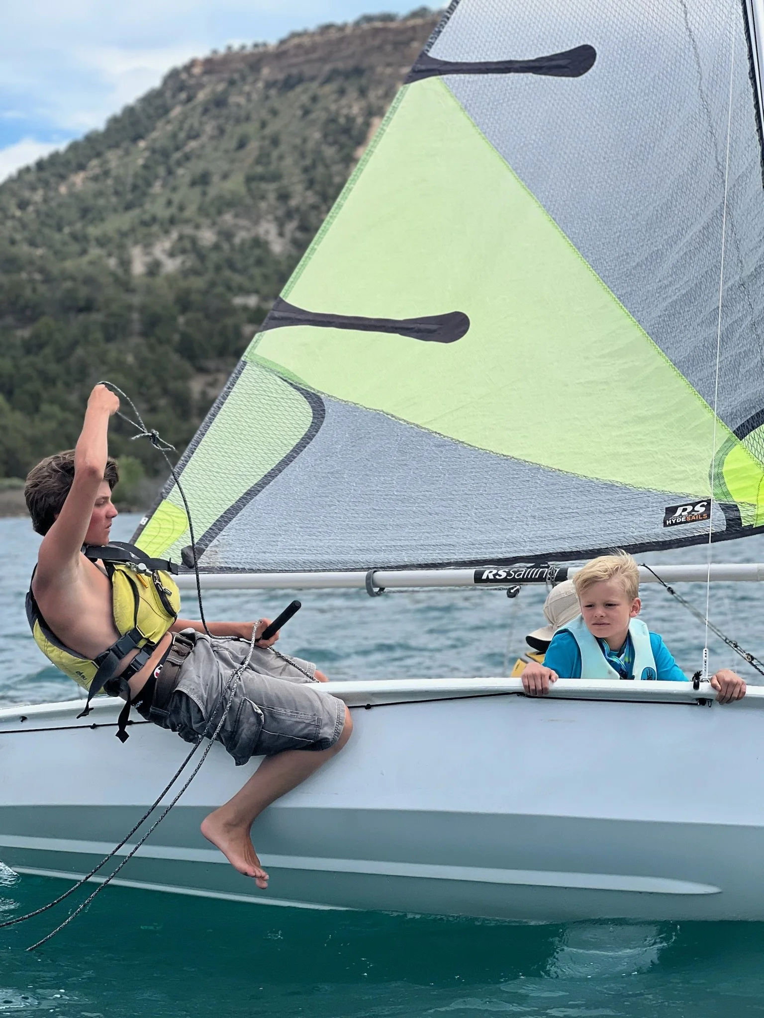 Two young boys are sailing a small boat on the water; one boy is sitting on the edge and pulling a rope, while the other boy is holding onto the side of the boat.