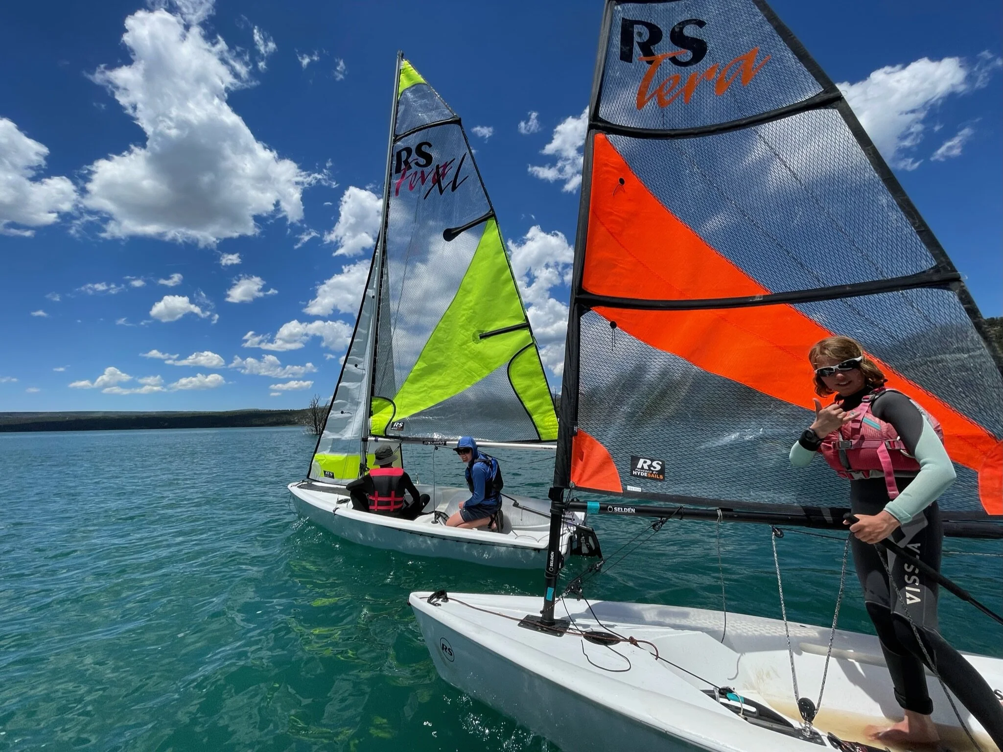 Three people sailing on small sailboats on a bright day with a blue sky and scattered clouds.