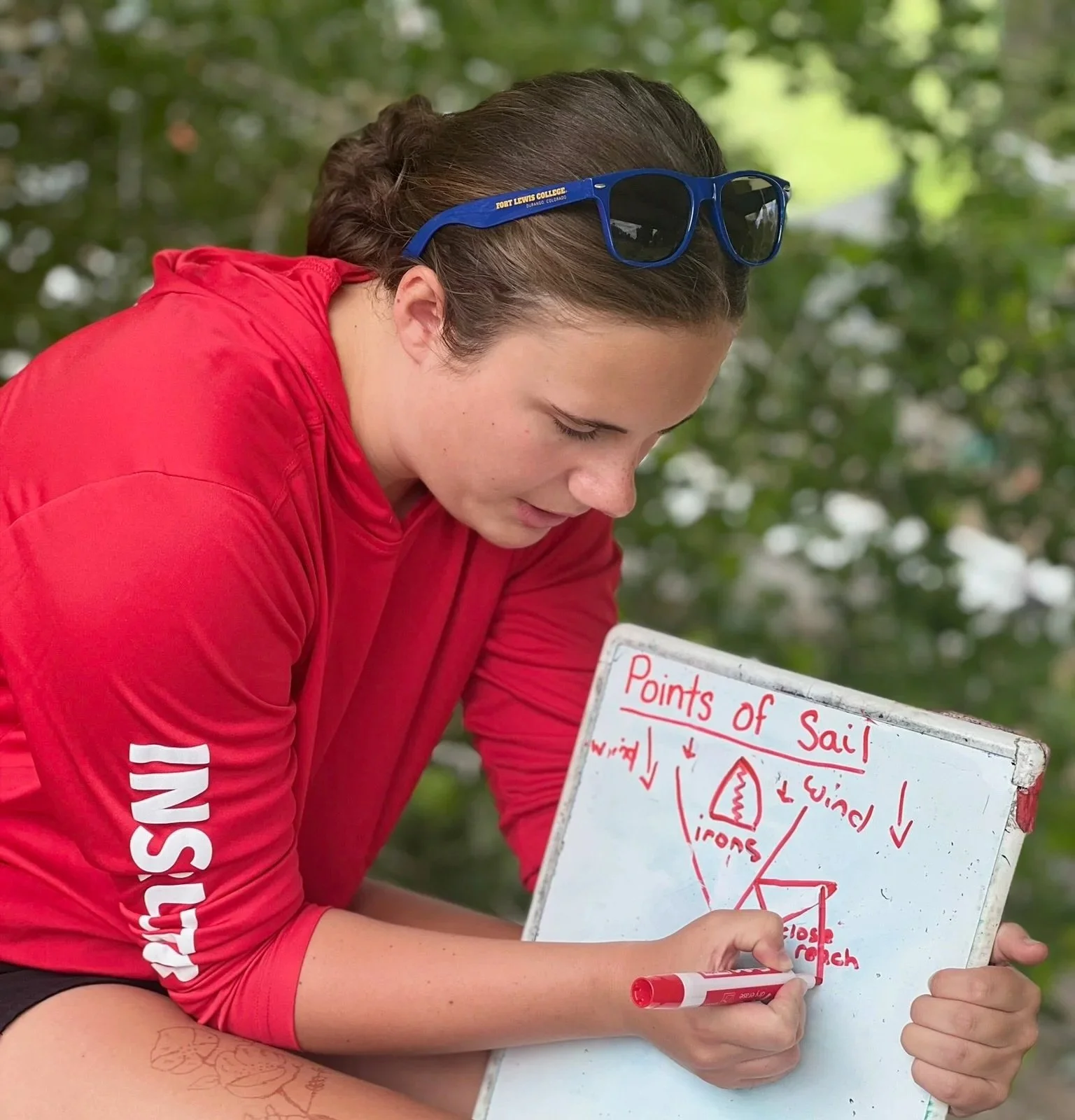 A person in a red shirt with 'INSY' printed on the sleeve is sitting outdoors, writing on a whiteboard with a red marker. The whiteboard has a diagram labeled 'Points of Sail' with arrows and labels about sailboat points and directions. The person is