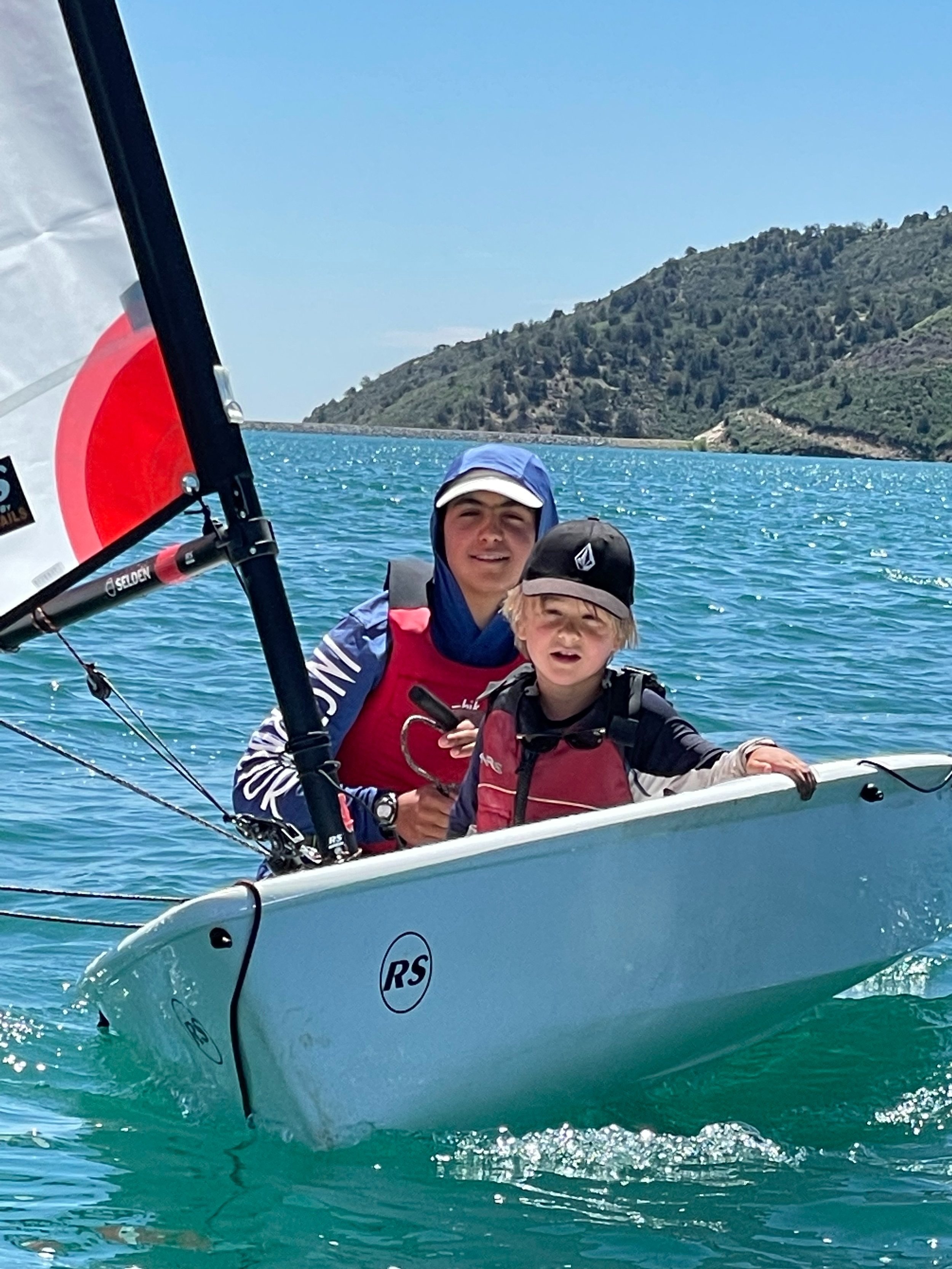 Two children sailing on a small sailboat on clear blue water with hills in the background, one wearing a blue jacket and cap, and the other in a red life vest and black cap.