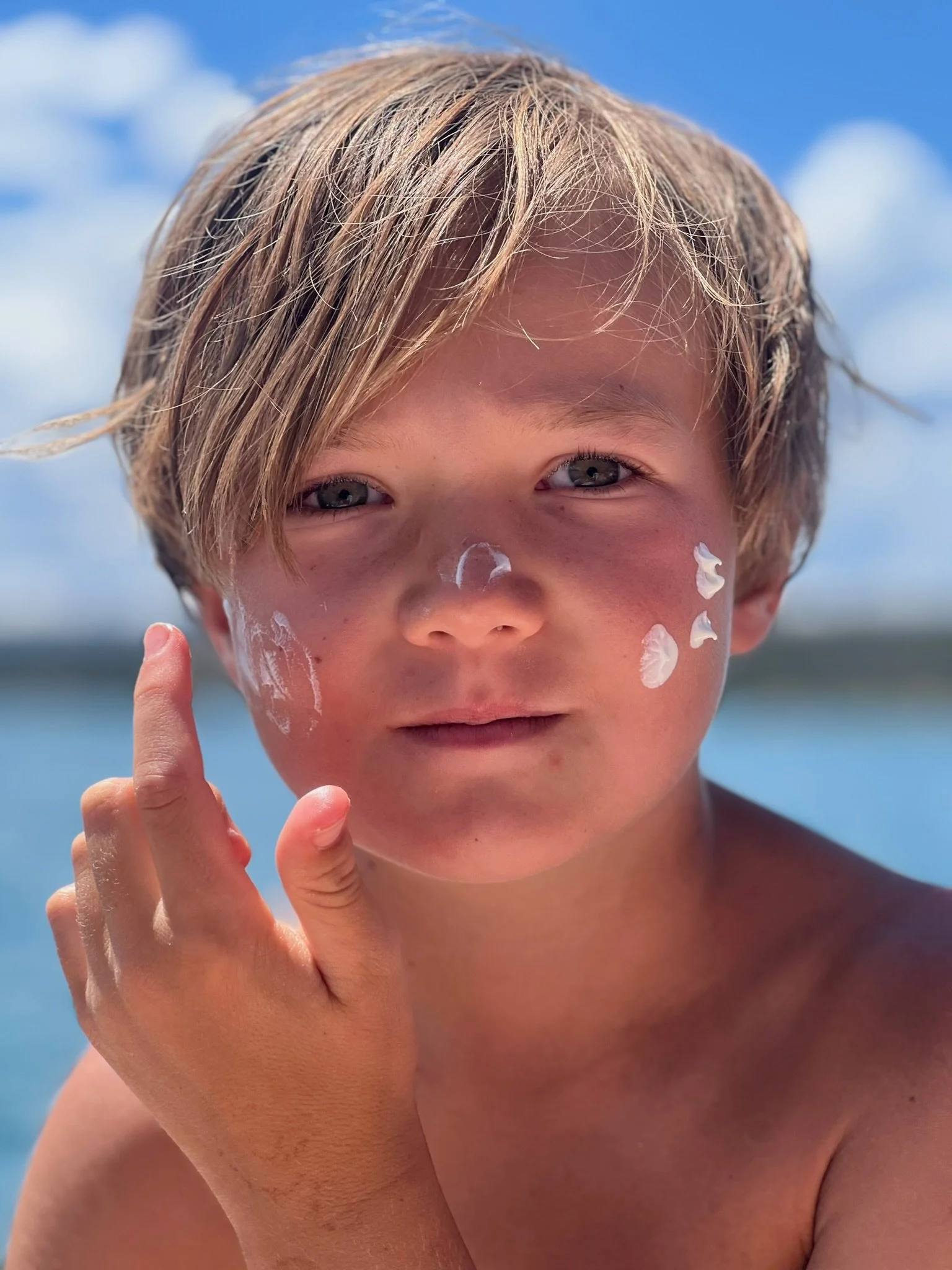 Young boy with wet hair outdoors, with paint on his face, touching his face, and a body of water in the background under a partly cloudy sky.