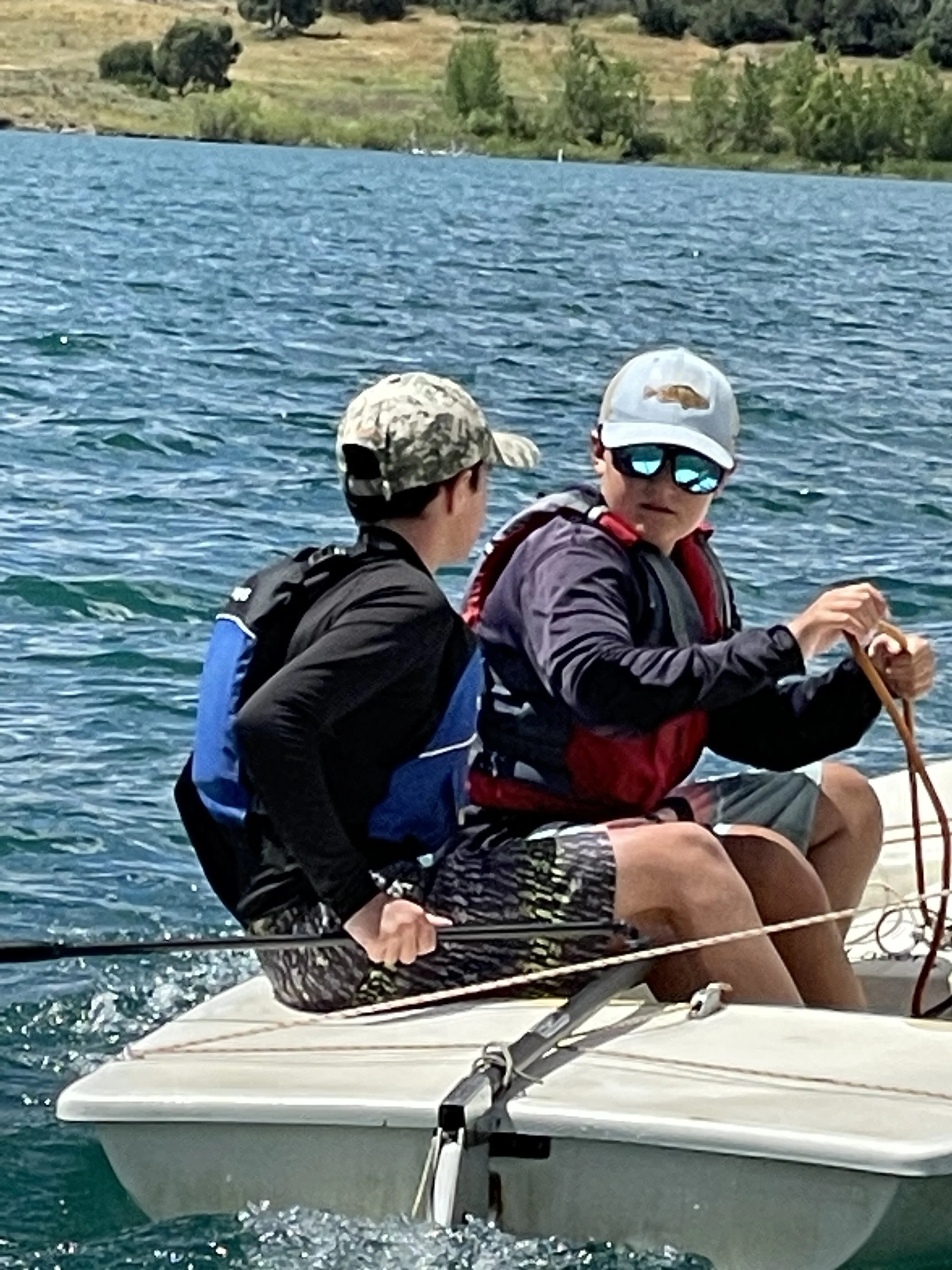 Two boys sitting on a small boat fishing on a lake, with hills and trees in the background, one wearing sunglasses and a cap.