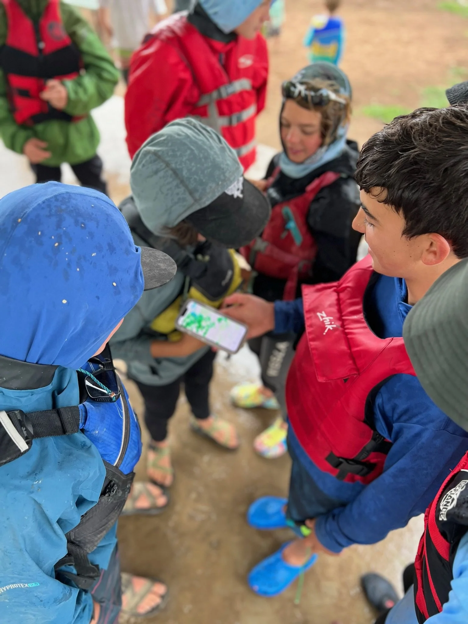 Group of children and teenagers dressed in outdoor gear and rain jackets, gathered in a circle outdoors, looking at a smartphone.