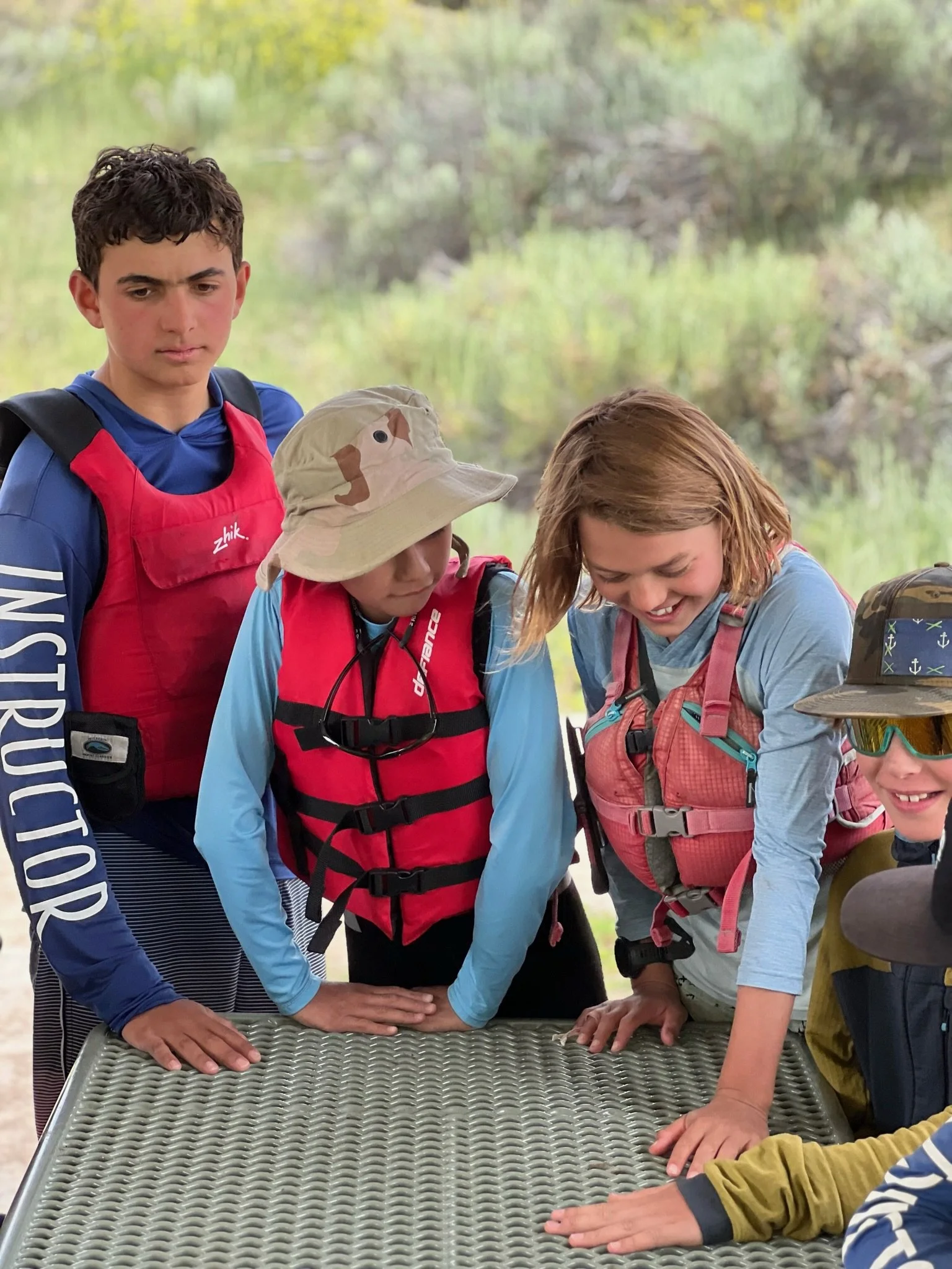 Group of children with backpacks gathered around a metal table outdoors, looking down at the table with interest and smiling.