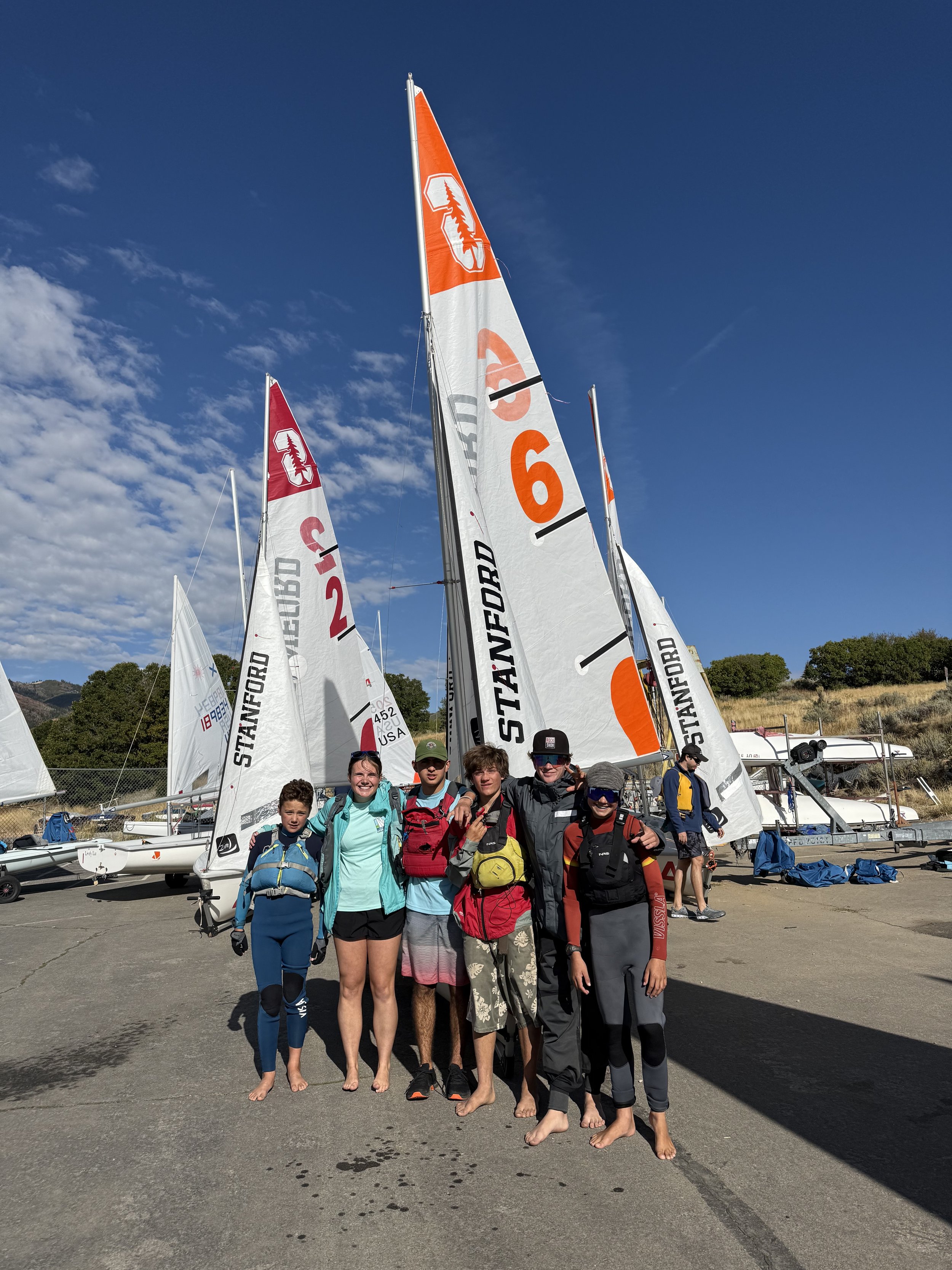 Group of people in sailing gear standing beside sailboats by a marina on a sunny day.