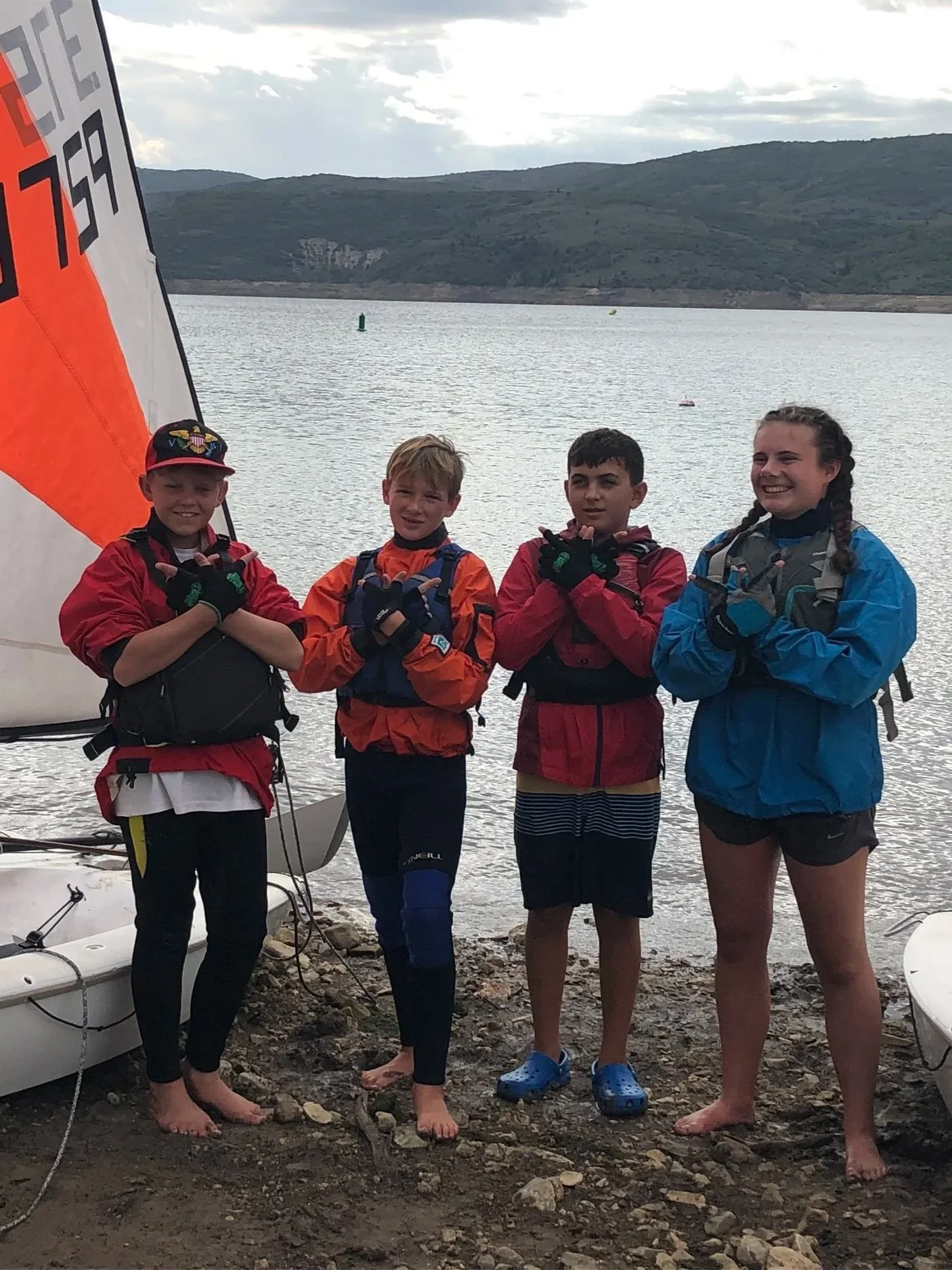 Four children standing barefoot on rocky shore near a lake, dressed in colorful outdoor jackets, with sailboats and hills in the background.