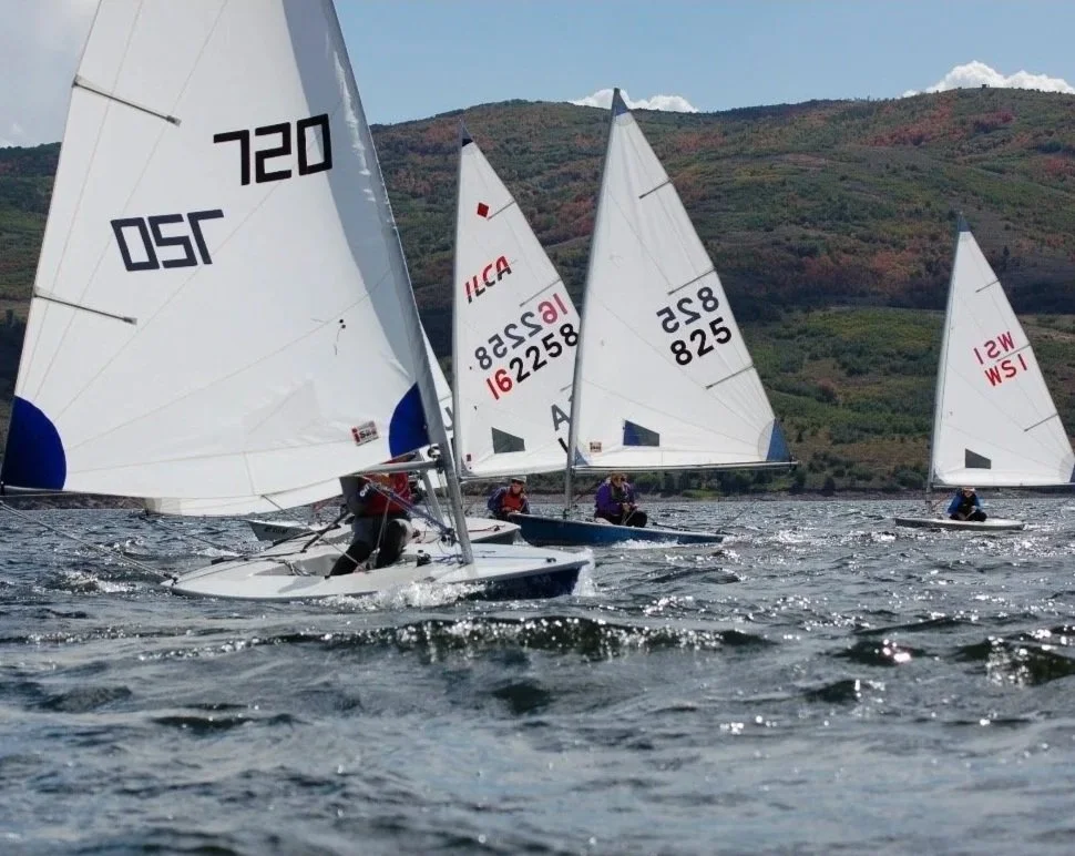 Multiple small sailboats racing on a lake with a hilly landscape in the background.