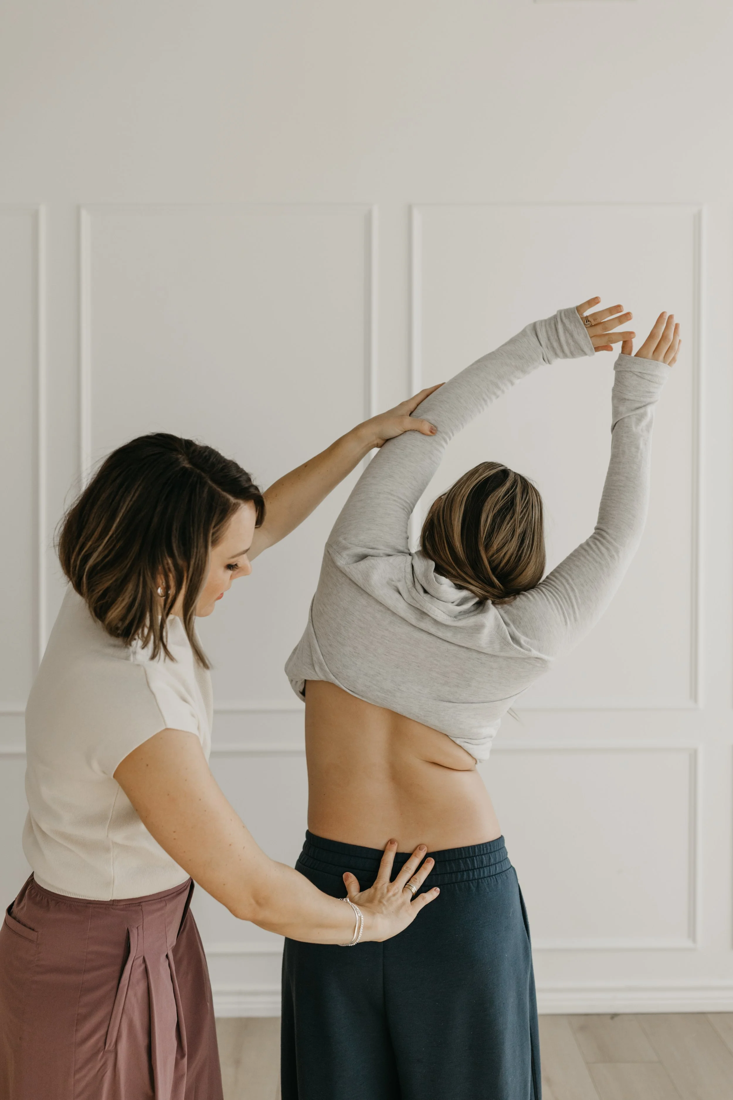 A woman is stretching her arms overhead while another woman assists her by placing her hands on her back and arm, in a room with white walls and wooden flooring.