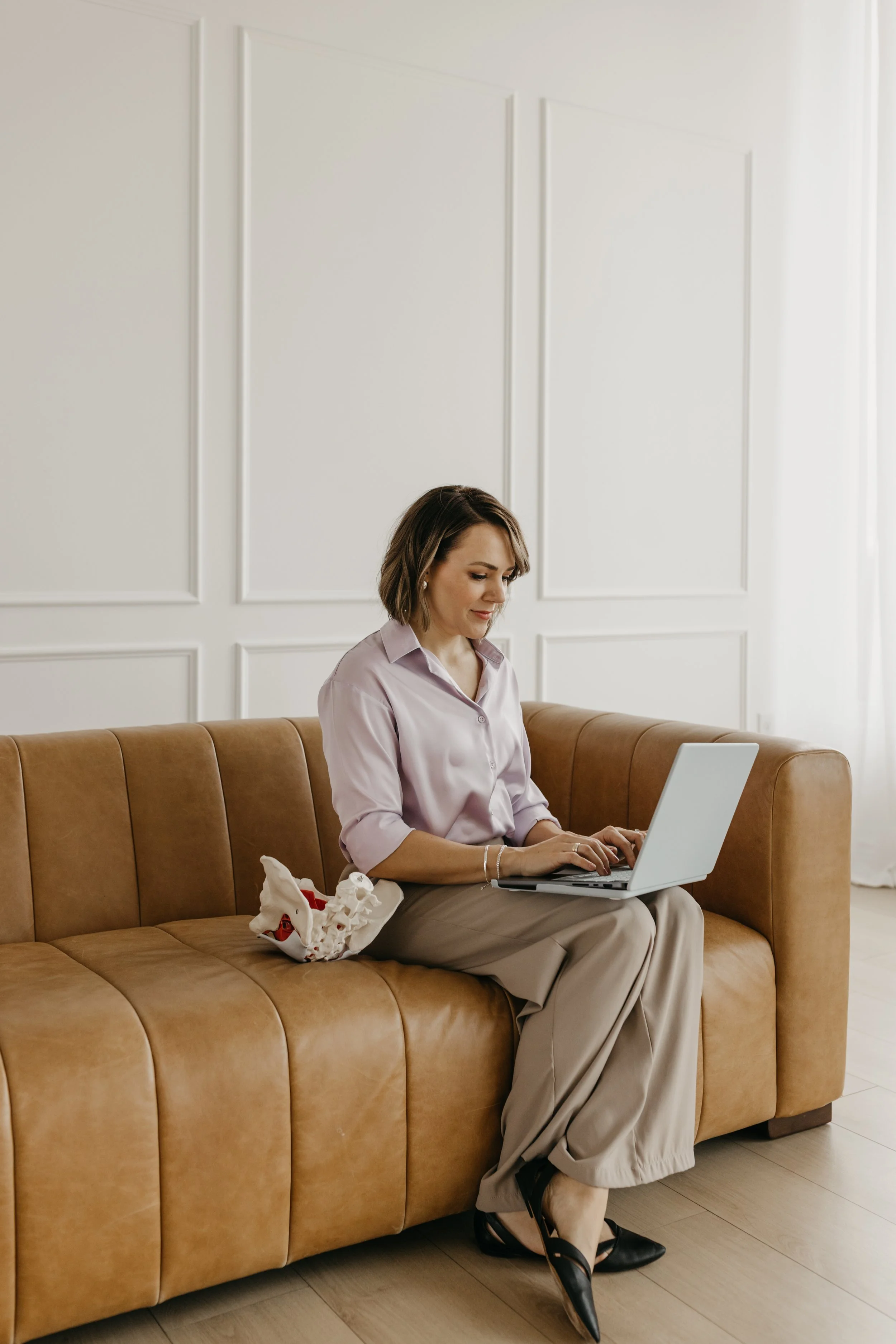 A woman with shoulder-length brown hair, wearing a light pink shirt and beige pants, sits on a tan leather sofa working on a silver laptop. Next to her on the sofa, there is a crumpled Christmas stocking with red and white colors. The room has white paneled walls and light wooden flooring.